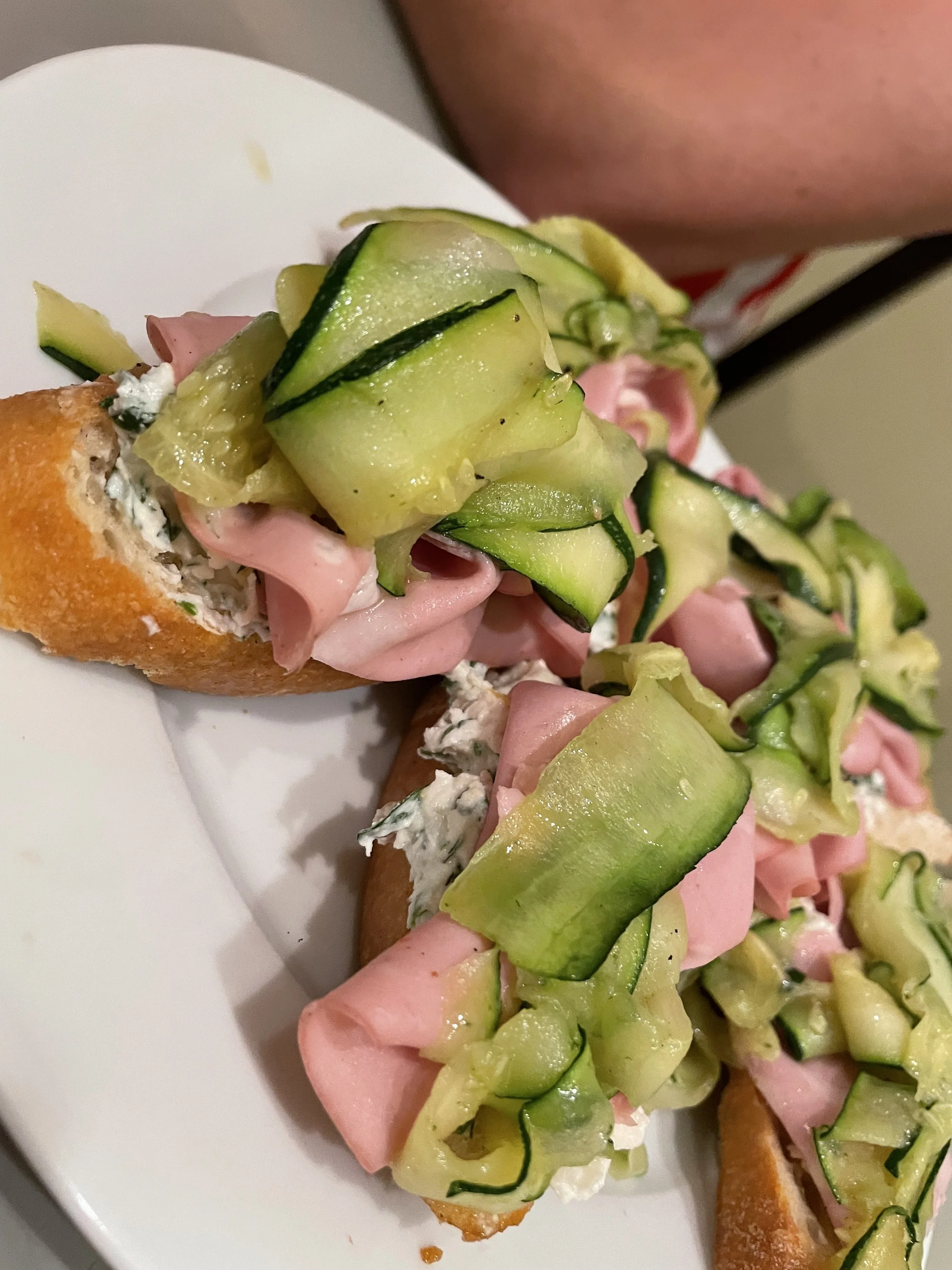 Close-up of open-faced sandwiches with sliced cucumbers, ham, and blue cheese spread on baguette slices.