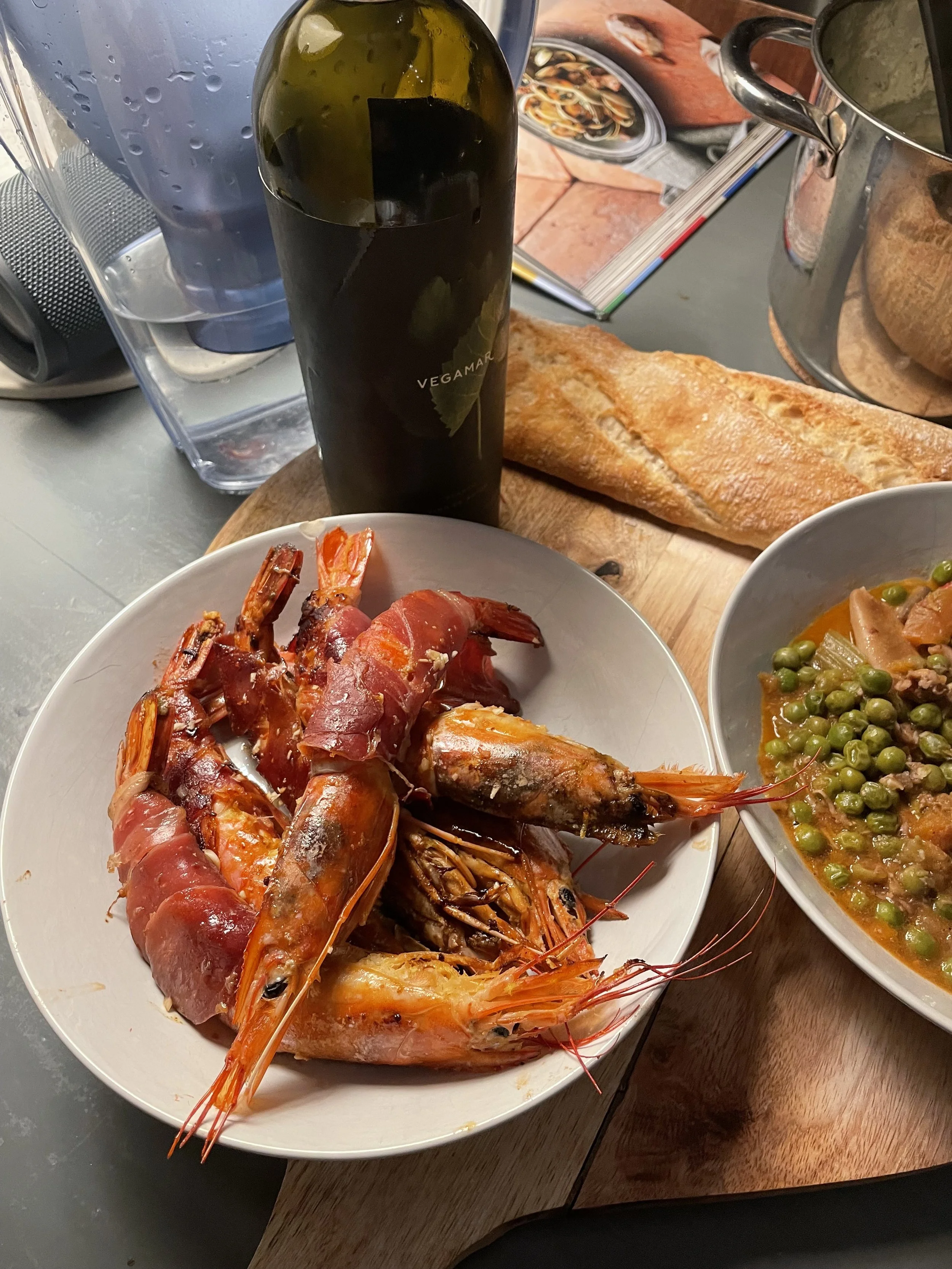 Plate of cooked prawns wrapped in prosciutto with a side bowl of green peas and vegetables, a bottle of wine, a loaf of bread, and an open cookbook on a table.