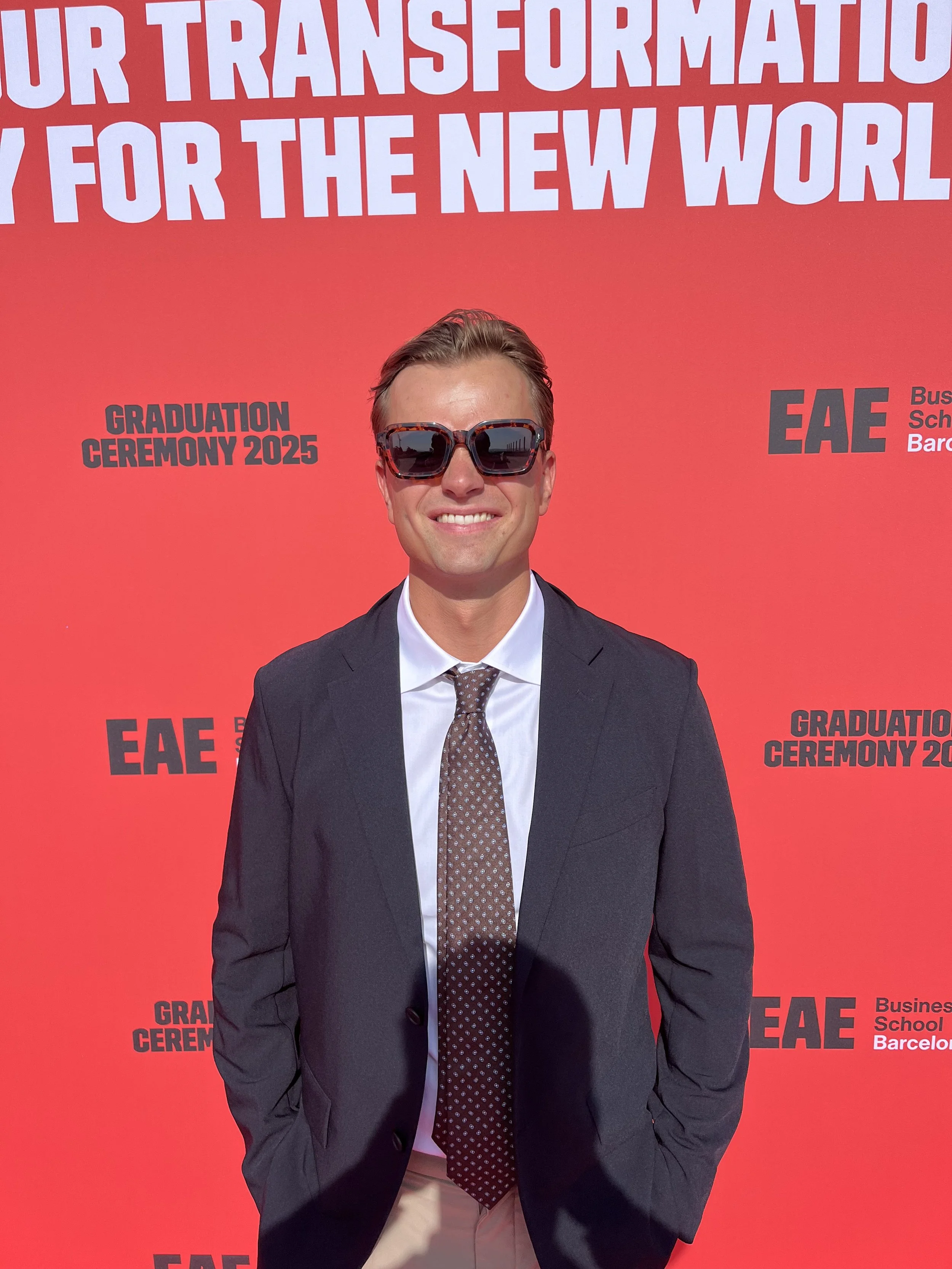 A young man wearing sunglasses, a white shirt, a dark blazer, and a patterned tie, smiling at a graduation ceremony in 2025. The background is red with white and black text related to the event.
