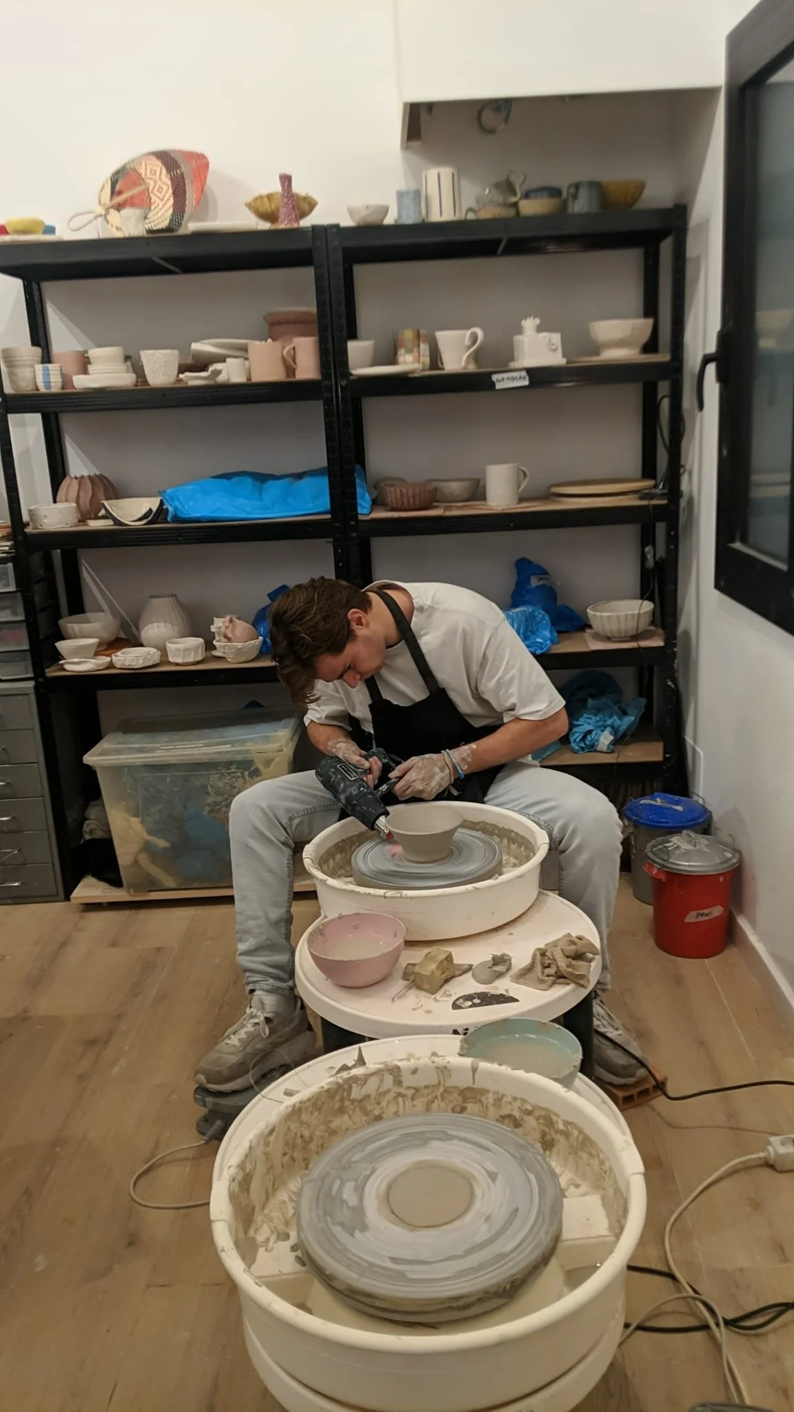 A person working on a pottery wheel in a ceramics studio, surrounded by shelves filled with various ceramic pieces and tools.