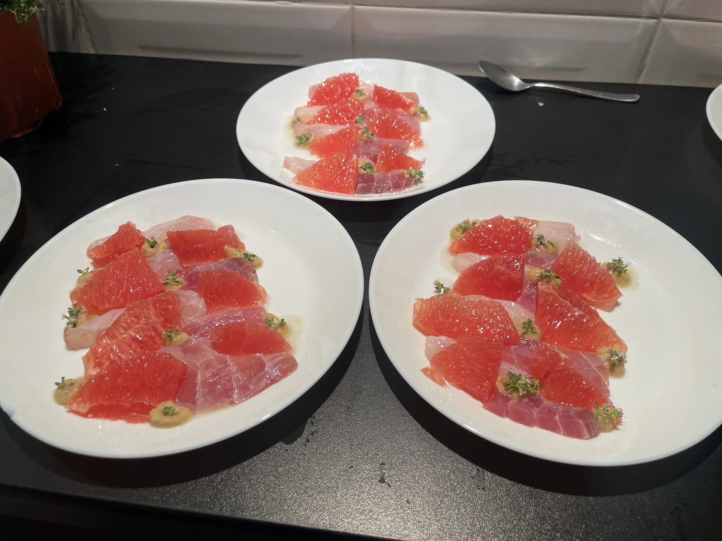 Three white plates of sliced raw fish, including salmon and possibly tuna, garnished with small herbs, on a black surface.
