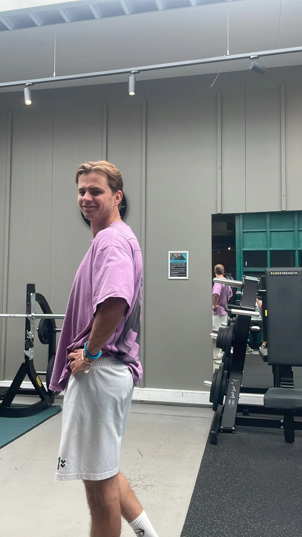 Young man with light brown hair wearing a purple t-shirt and white shorts standing inside a gym, with gym equipment and a mirror in the background.