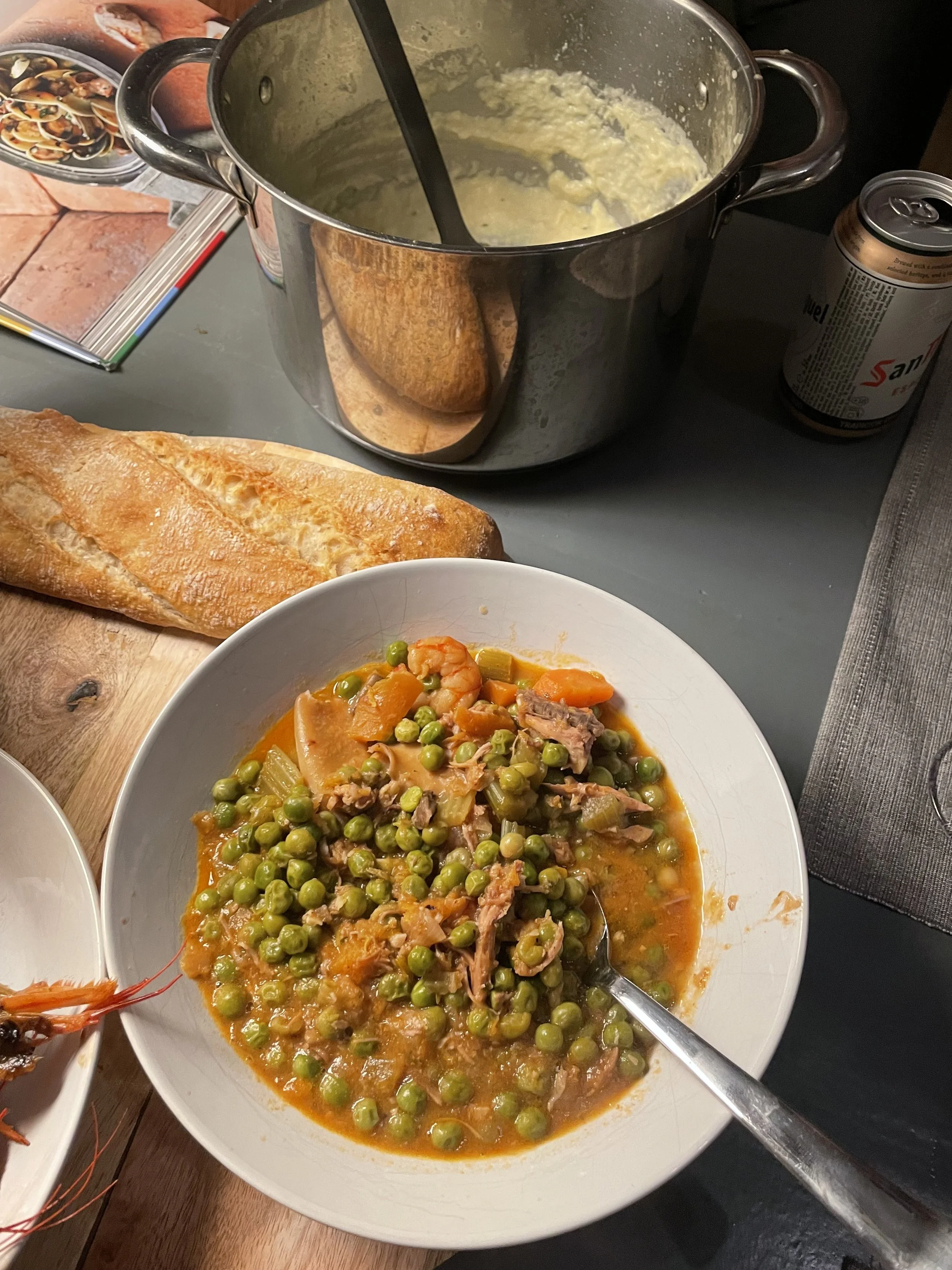 A bowl of vegetable stew with green peas, carrots, and meat, with a spoon inside, on a dining table. Nearby, a loaf of baguette bread. In the background, a large pot with mashed potatoes or similar dish, a can of San Pellegrino sparkling water, and a