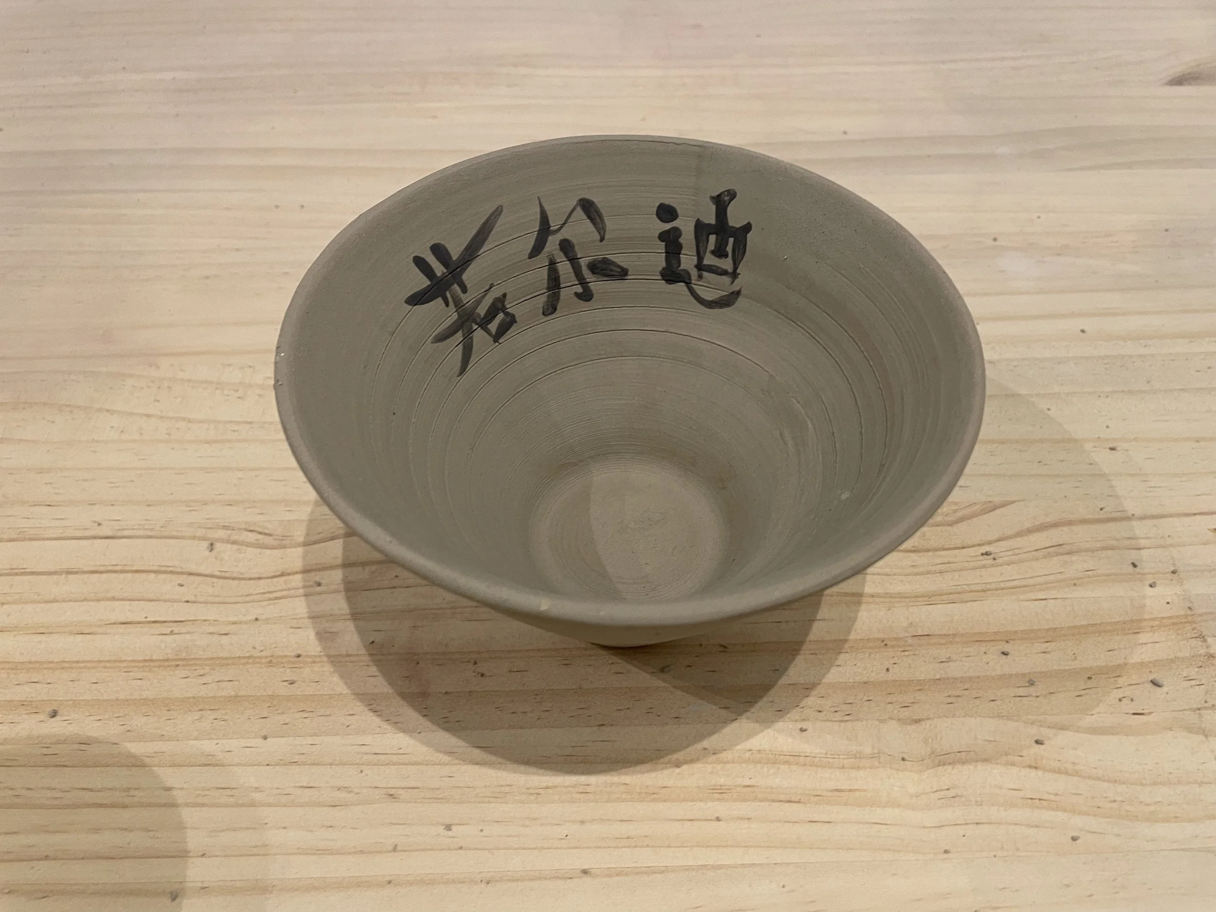 Empty ceramic bowl with black Chinese characters written on the inside, placed on a light wooden surface.