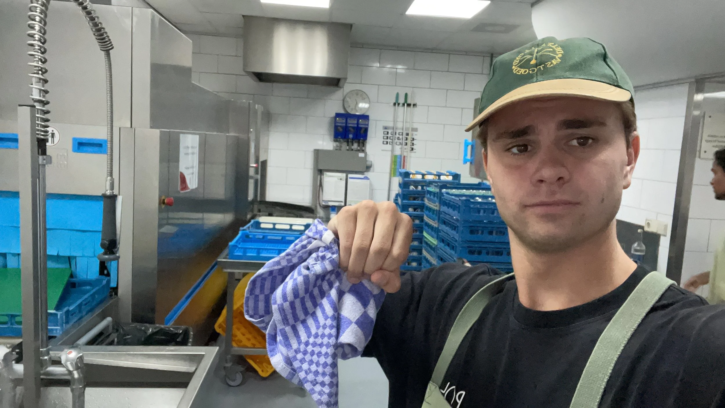 A young man taking a selfie inside a commercial kitchen, holding a blue and white checkered dishcloth. He is wearing a green cap with yellow writing, a black T-shirt, and a light green apron straps. The background includes stainless steel equipment, 