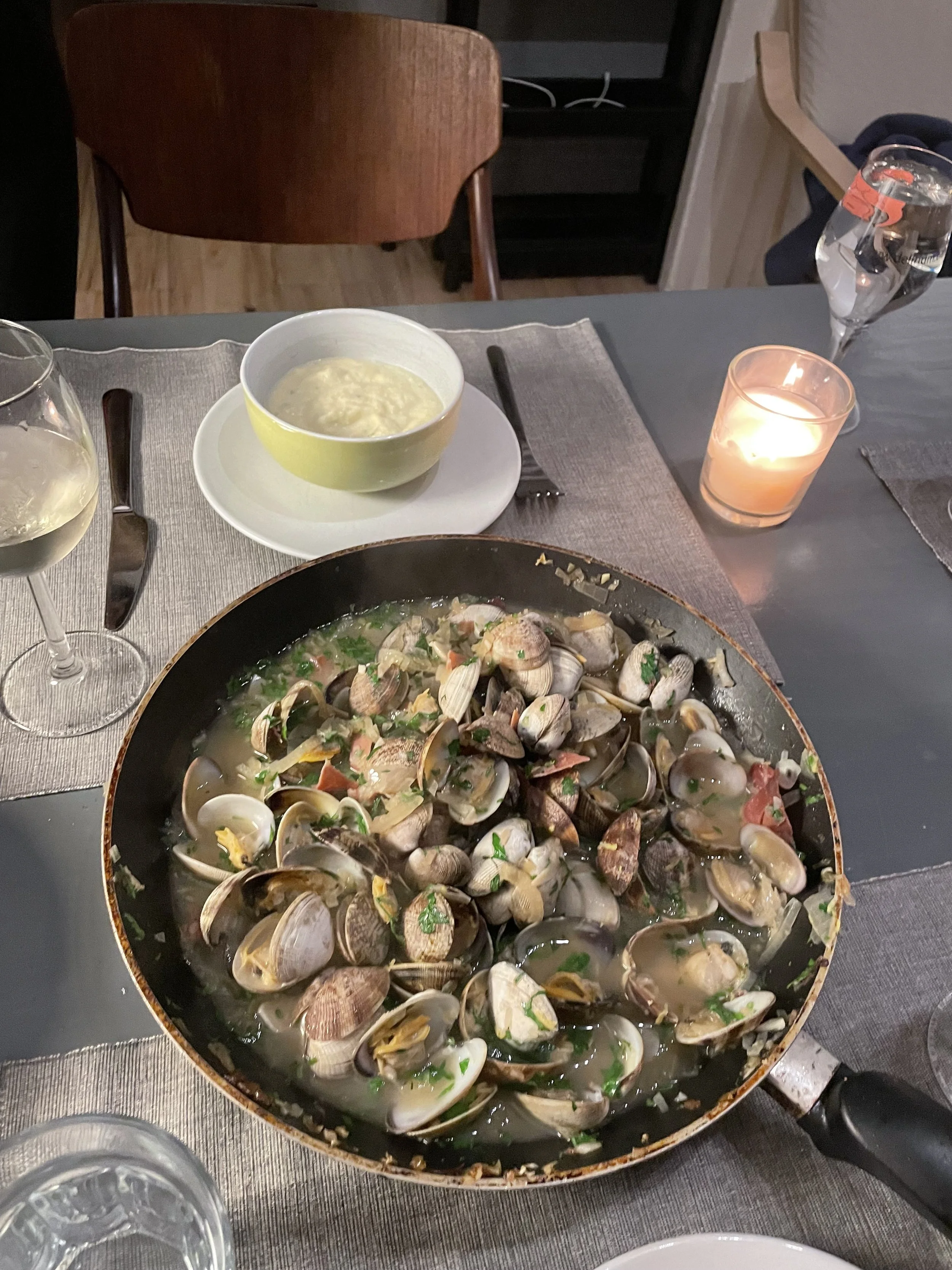 A table set for dining with a large pan filled with clams in broth, a bowl of mashed potatoes, two glasses of white wine, a lit candle, and a glass of water.