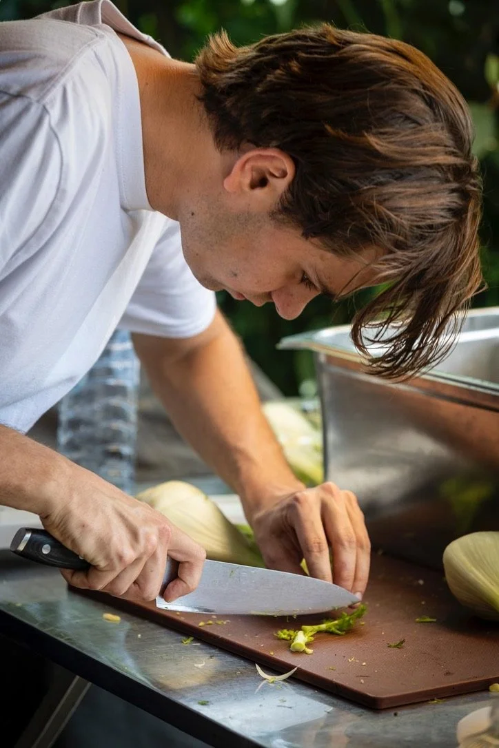 Young man chopping green onion with a large knife on a brown cutting board, with fennel bulbs nearby, outdoors.