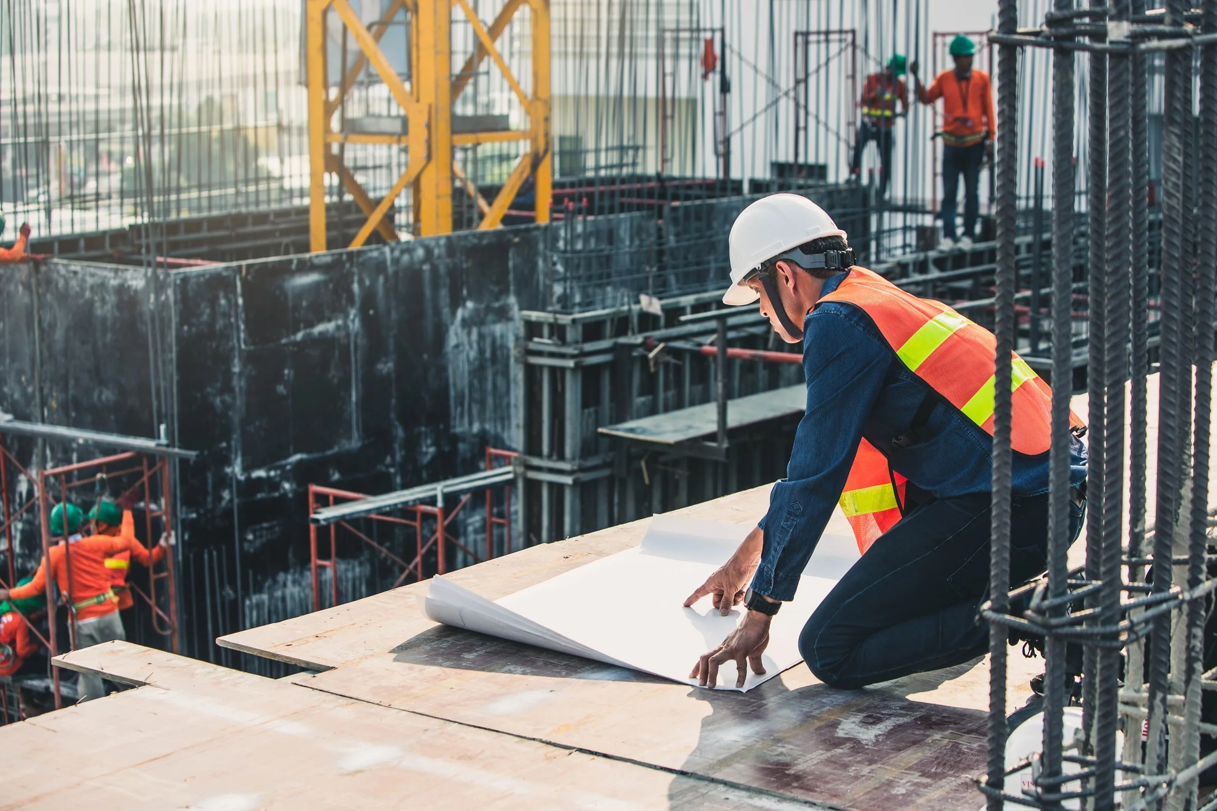 LYS (Lai Yew Seng)-services-A construction worker kneeling on a platform, looking at blueprints on a table, wearing a white helmet and safety vest, with a construction site with scaffolding and workers in the background.