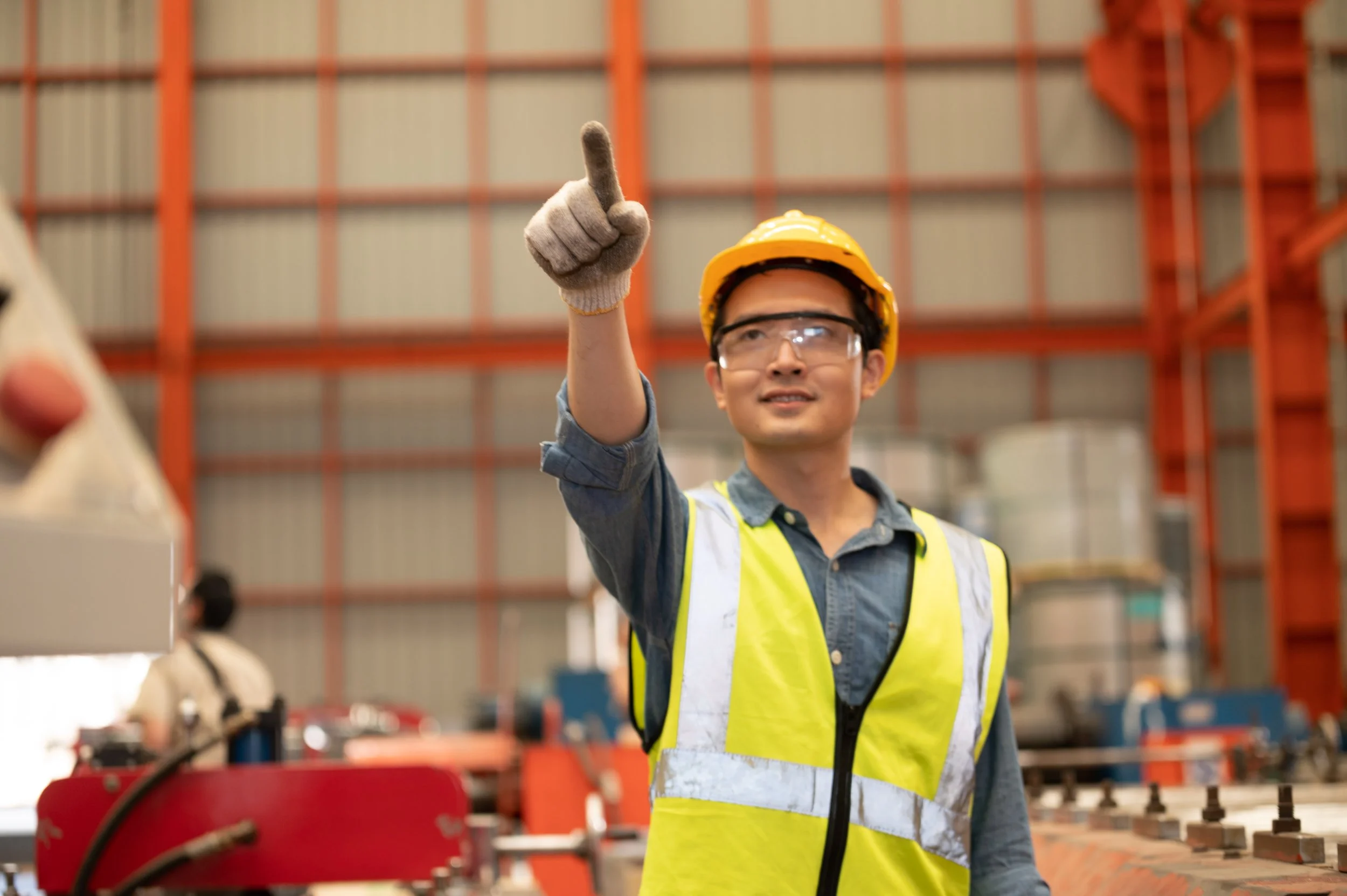 A young male construction worker wearing a yellow safety helmet, clear safety glasses, and a bright yellow safety vest pointing upward inside a warehouse or factory.