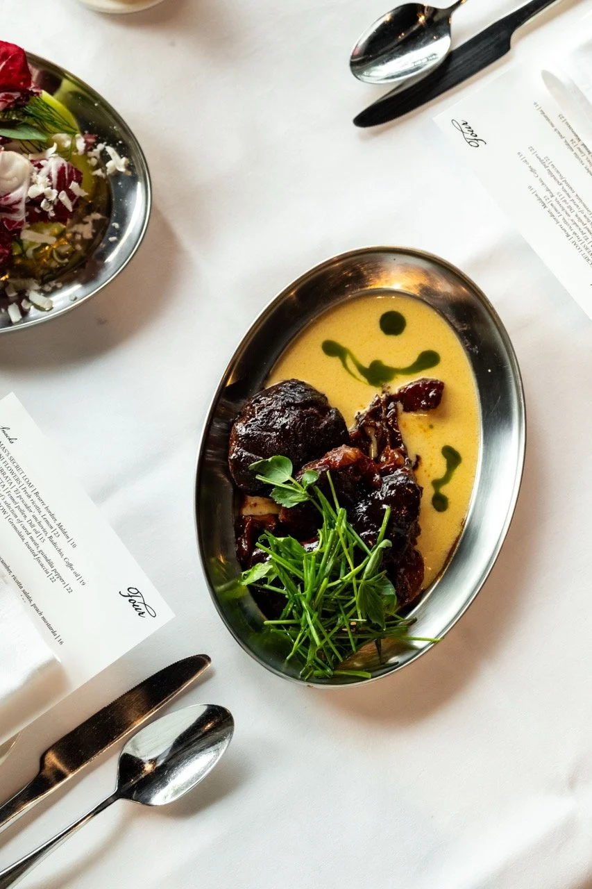 A metal dish containing a serving of braised beef with green herbs, surrounded by creamy yellow sauce with green oil drops, on a white table with utensils