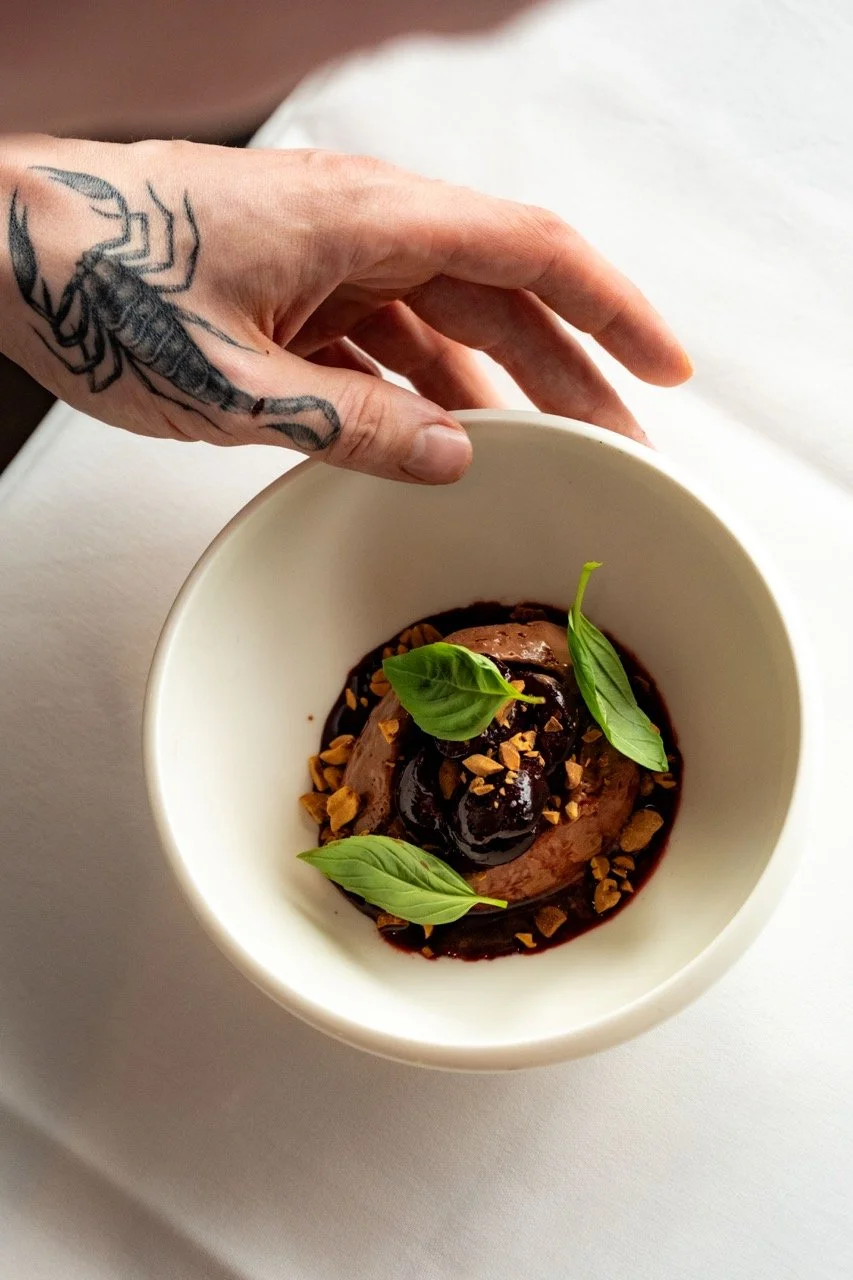 A hand with a tattoo of a scorpion holding a white bowl containing a dessert topped with chocolate sauce, chopped nuts, and fresh basil leaves.