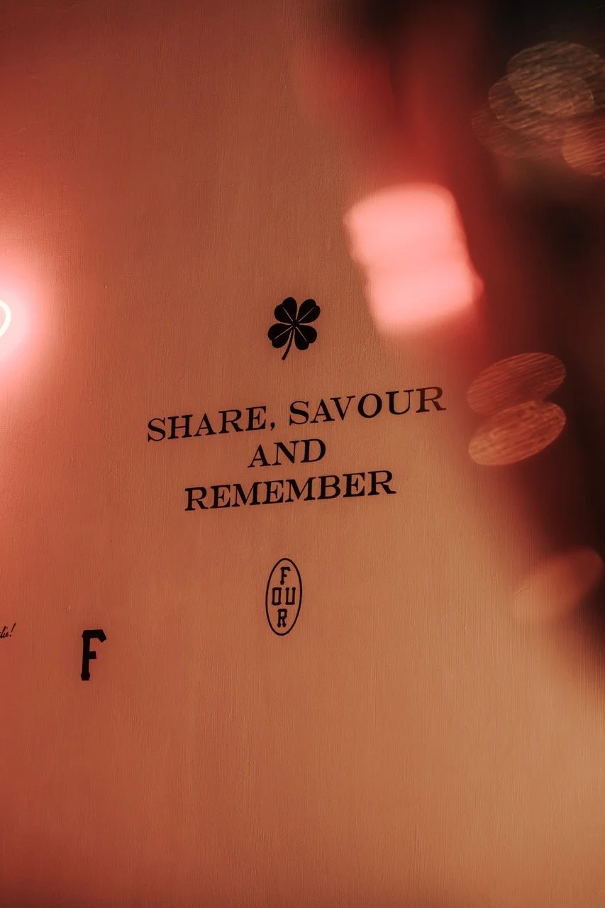 Close-up of a sign with a four-leaf clover symbol and the words 'Share, Savour and Remember' in black text, in a warm, pinkish lighting environment.