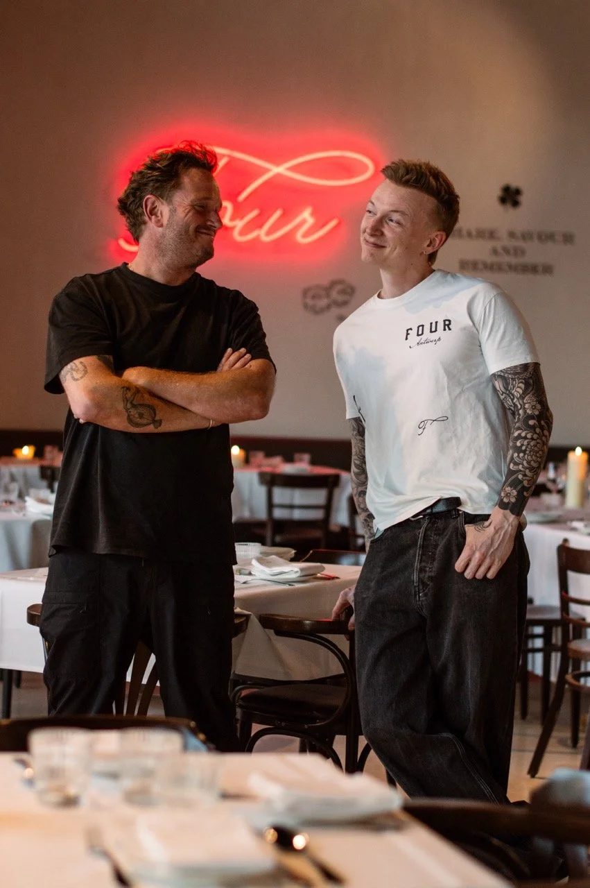 Two men in a restaurant, smiling and talking to each other. One man is wearing a black shirt with tattoos on his arms, and the other is wearing a white T-shirt with tattoos on his arms. There is a neon sign behind them that says 'Lair' and tables with candles in the background.