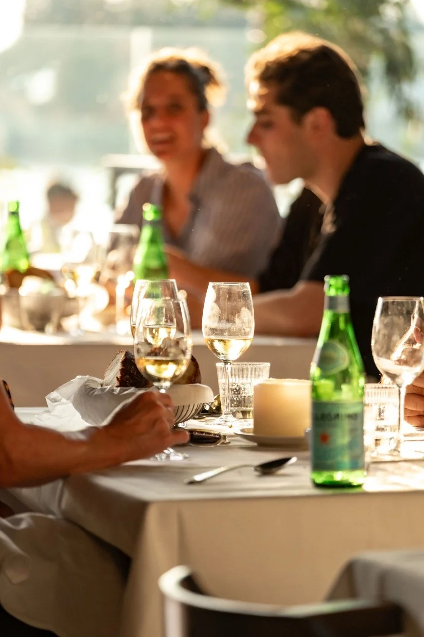 People sitting at a dining table with drinks and food, in a bright room with sunlight streaming in.