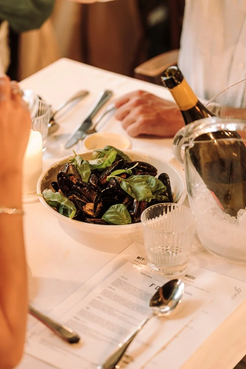 A table with a bowl of cooked mussels topped with fresh basil, a glass of bottled beverage in an ice bucket, a glass of water, a spoon, a fork, and a menu on a white tablecloth.