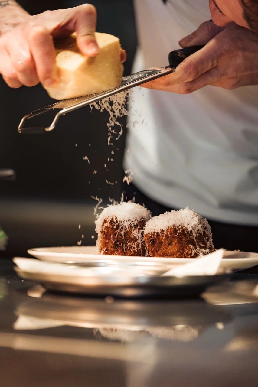 Close-up of a chef grating cheese over two fried food balls on a plate in a kitchen.
