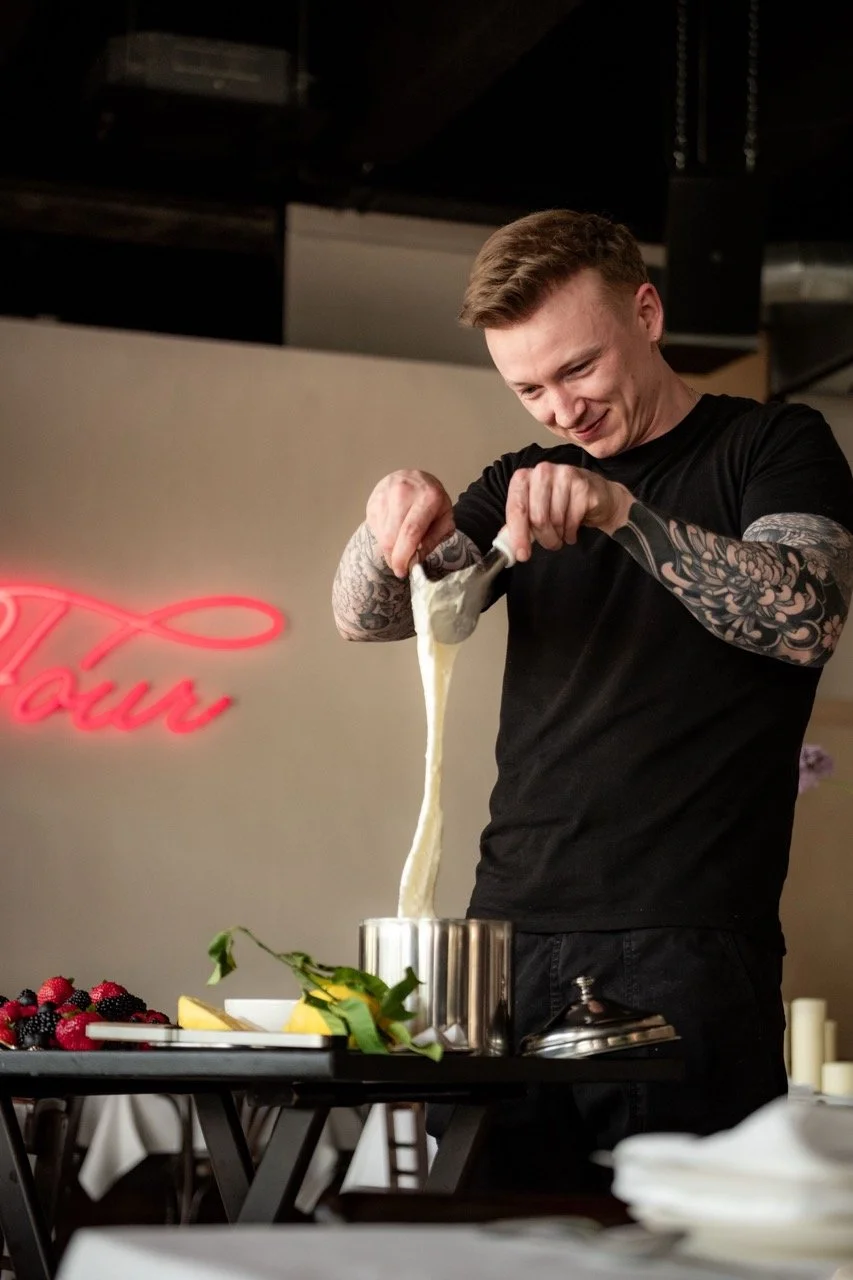 A man with tattoos on his arms, wearing a black T-shirt, is stretching mozzarella cheese over a pot in a restaurant kitchen. On the table in front of him are fresh berries and lemon slices.