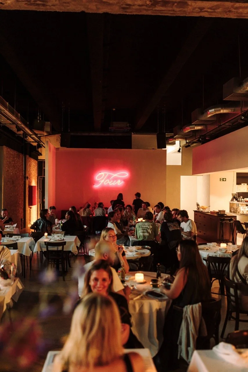 A busy restaurant filled with moonlit candlelit tables and patrons dining, with a neon pink sign reading 'Tour' on the wall.