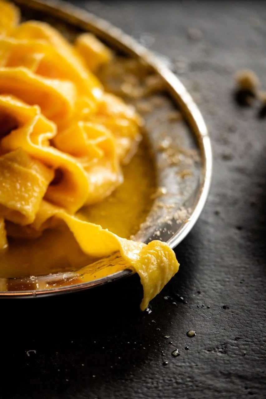 Close-up of cooked egg noodles in a metal bowl, with some noodles spilling onto a dark surface with a few black peppercorns.