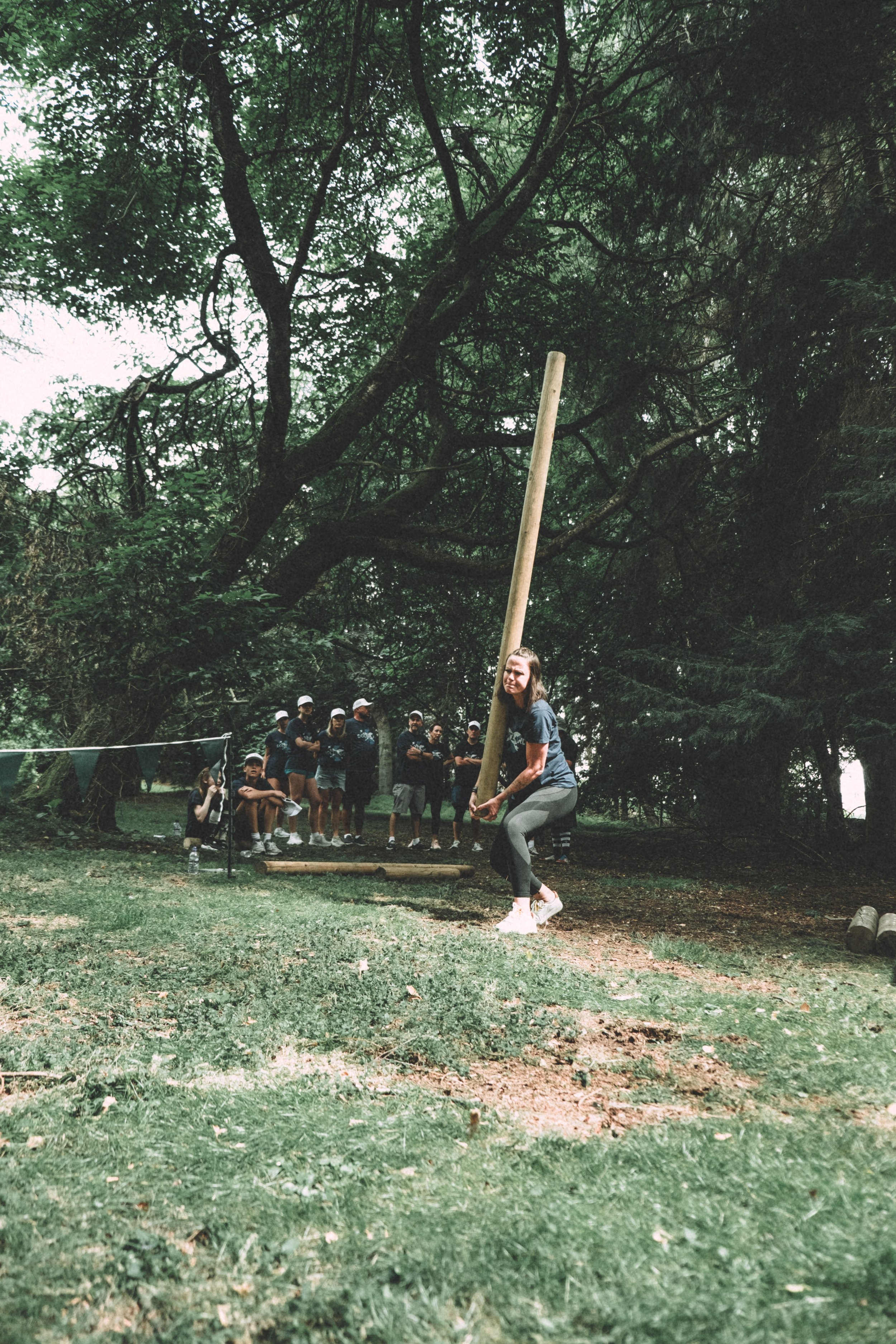 A woman is participating in a tug-of-war game outdoors on a grassy area with trees overhead. She is pulling on a long wooden pole, and a group of people is standing in the background, watching the activity.