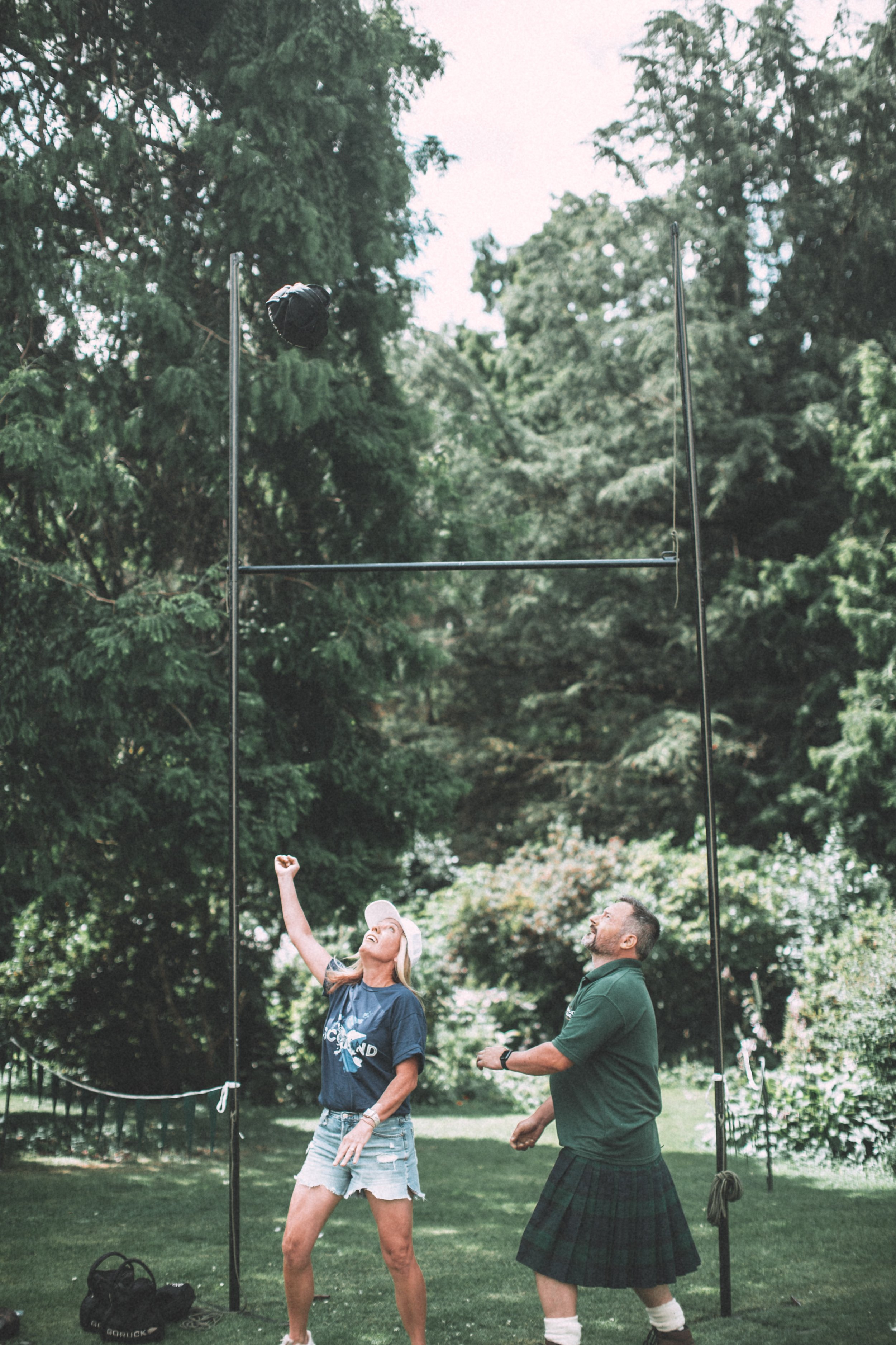Two people standing outdoors in front of a pole vault setup, with a woman raising her arm and a man dressed in a kilt looking on, surrounded by trees.