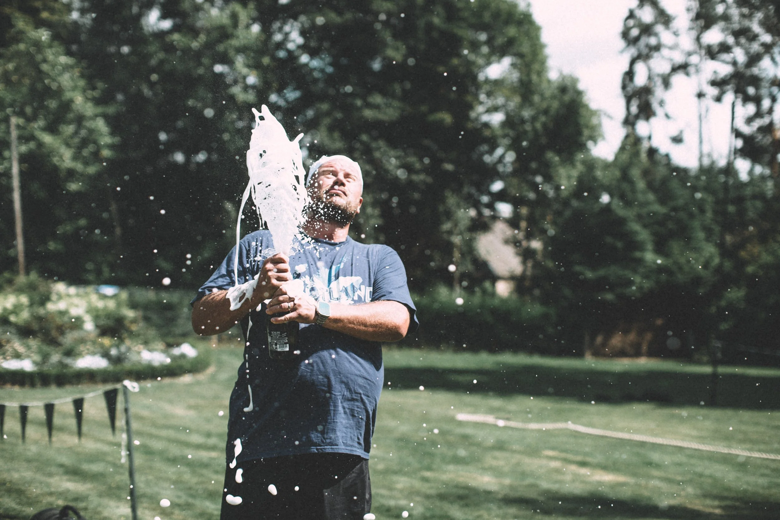A man with a beard and wearing a blue t-shirt is splashing water on himself outdoors in a park with green trees in the background.