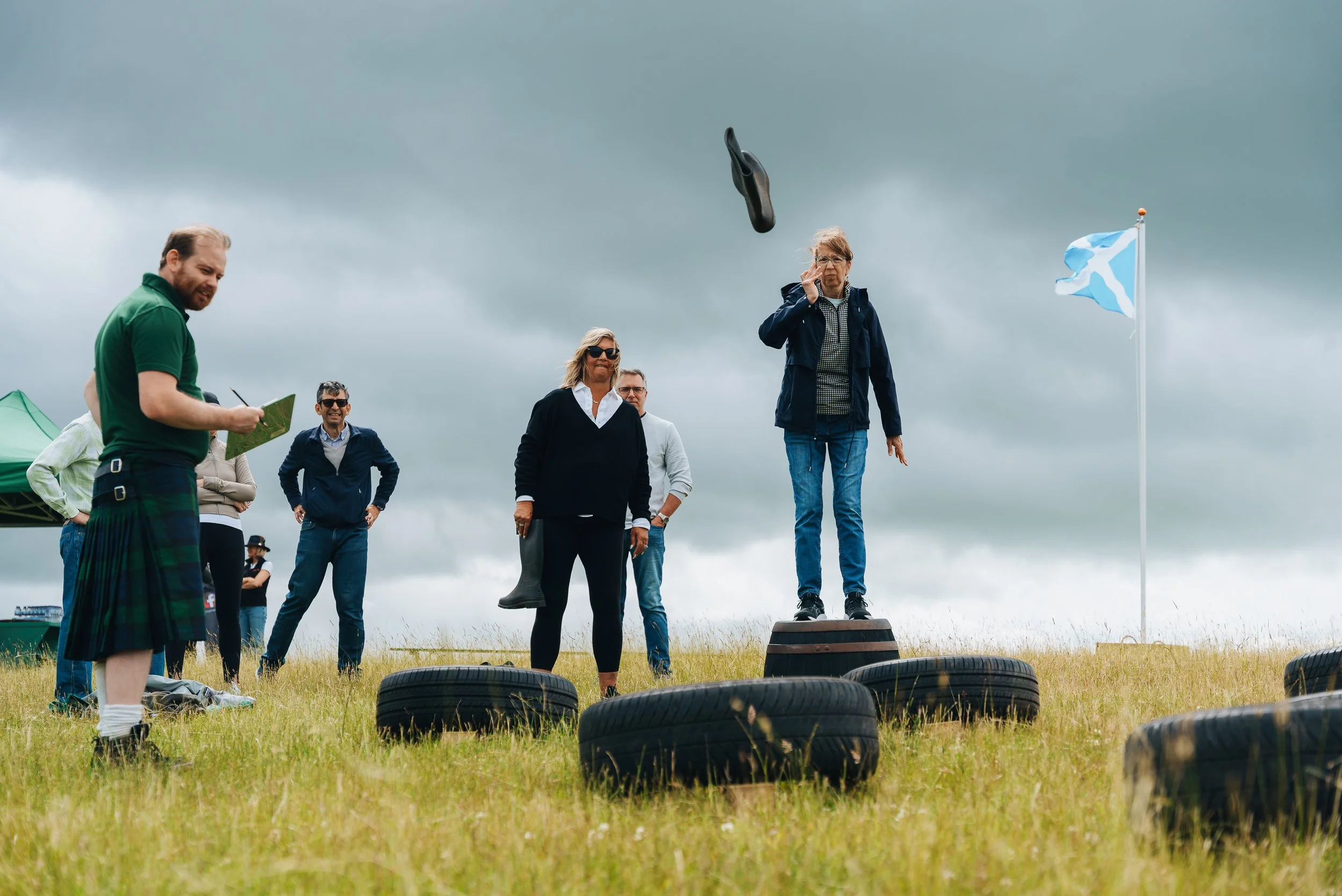 A group of people watching a woman toss a black shoe into the air at an outdoor event on a cloudy day, with a flag and tires on the grass.