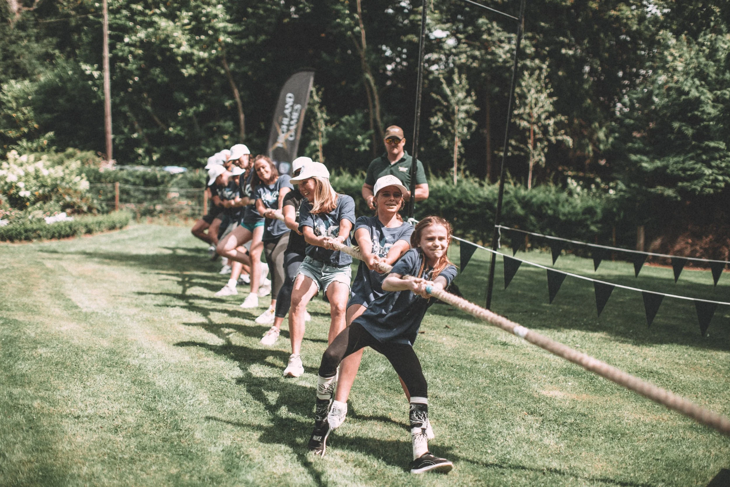 Children and adults participating in a tug-of-war game outdoors on a grassy field surrounded by trees, with some spectators and volunteers watching.
