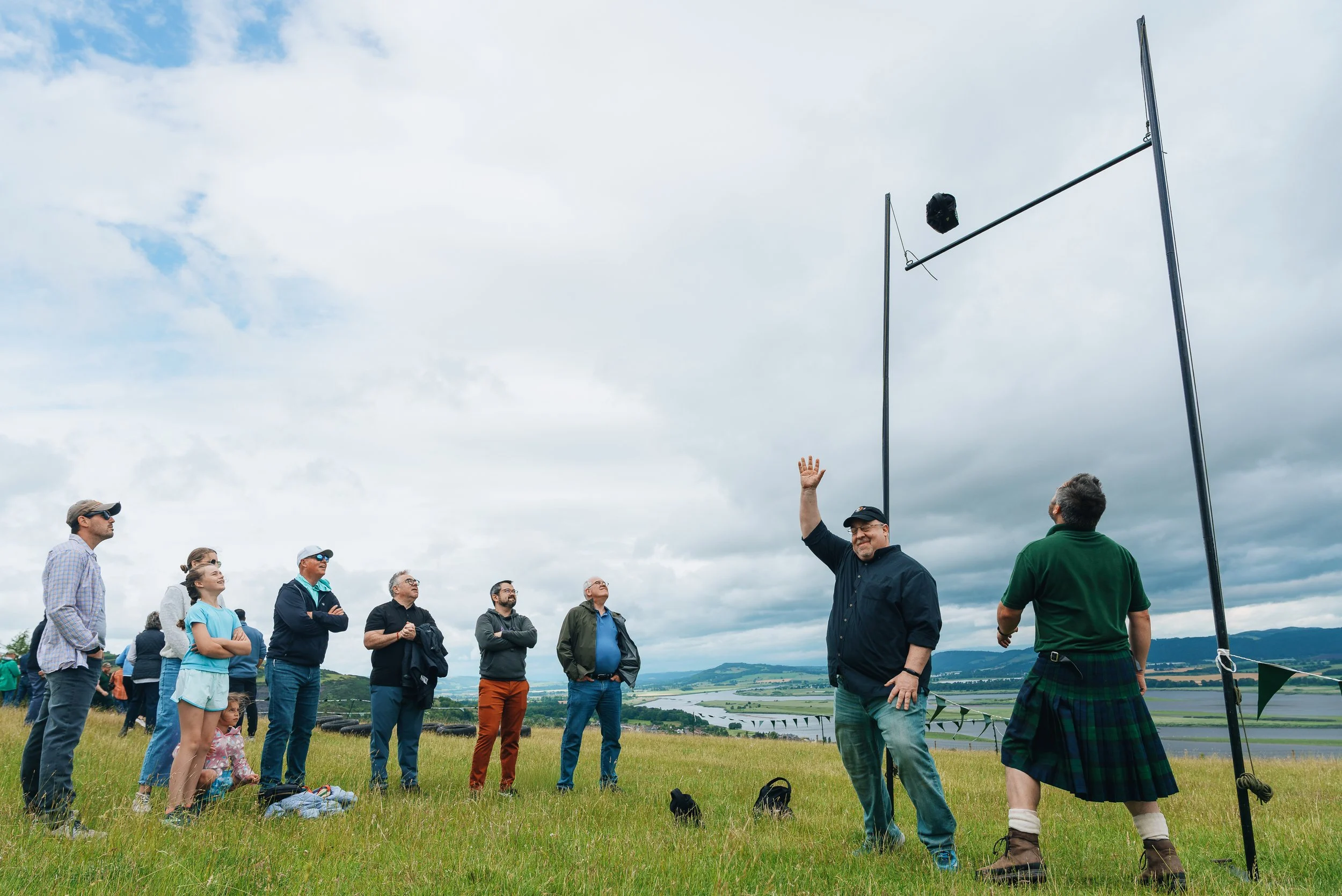 A group of people watching a man launch a model rocket outdoors on a grassy field with a cloudy sky and a scenic landscape in the background.