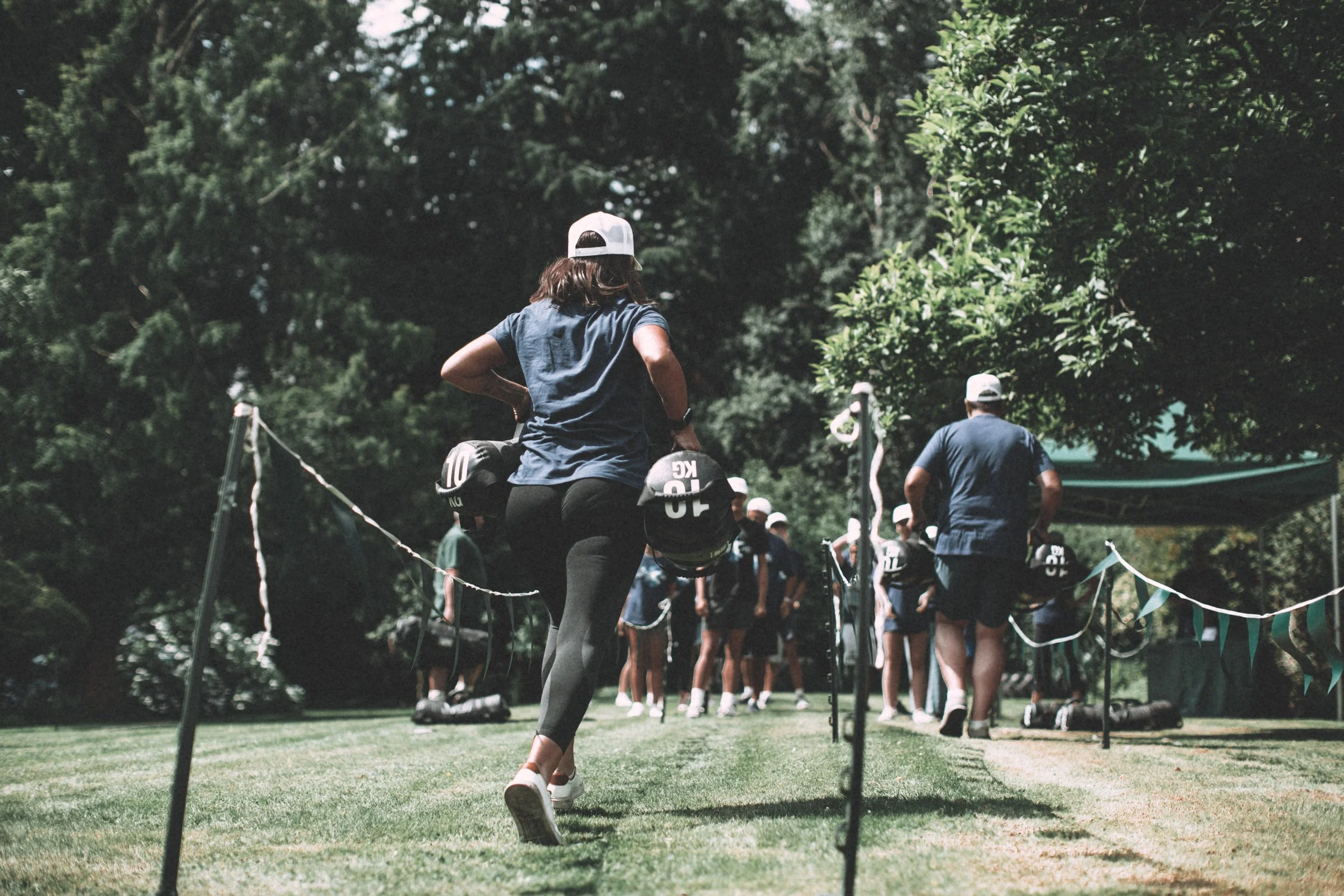 A group of children and adults participating in an outdoor activity in a park, with trees and green foliage in the background. Some children are wearing helmets, and one woman is seen from behind wearing a white cap and denim shirt.