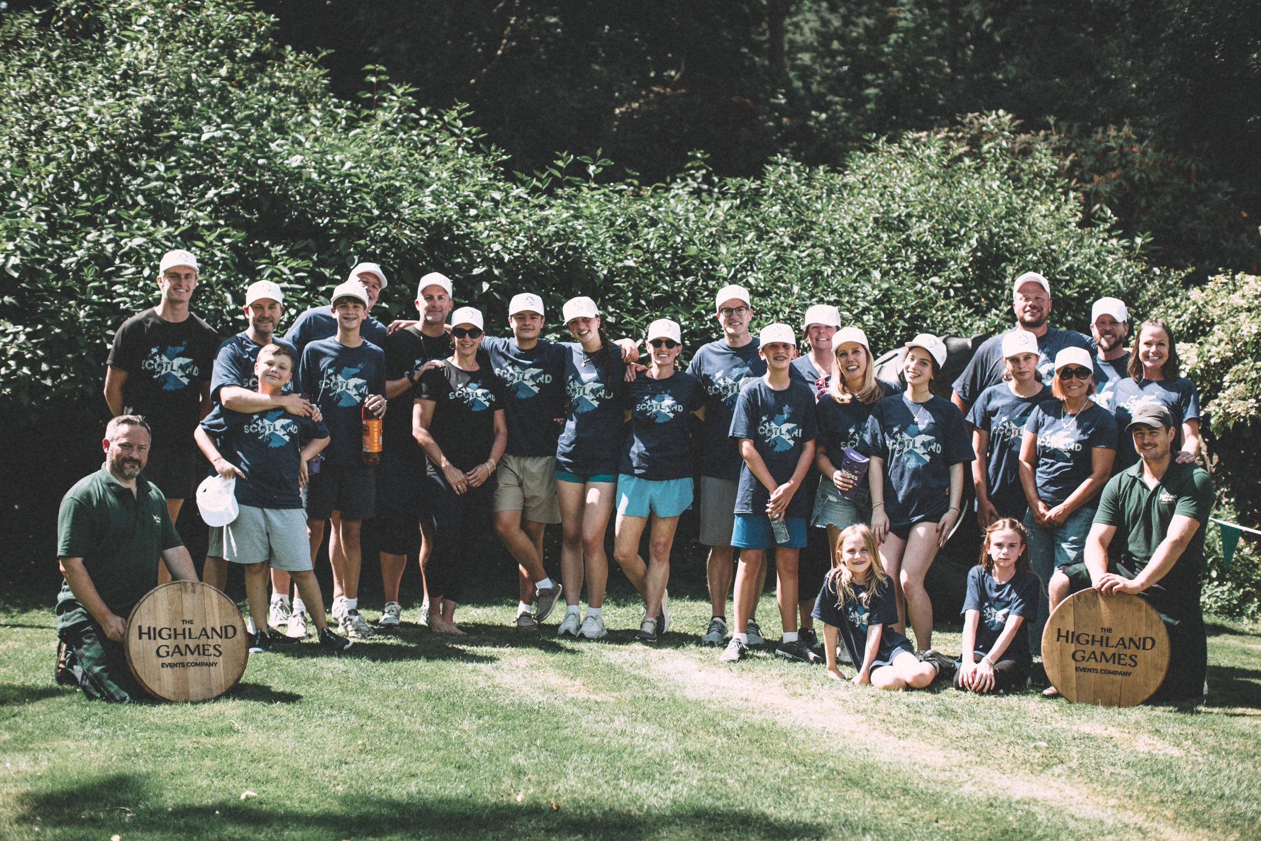 Group of people at Highland Games event, standing outdoors on grass with trees in background, some wearing matching blue t-shirts and white hats, posing for a photo.