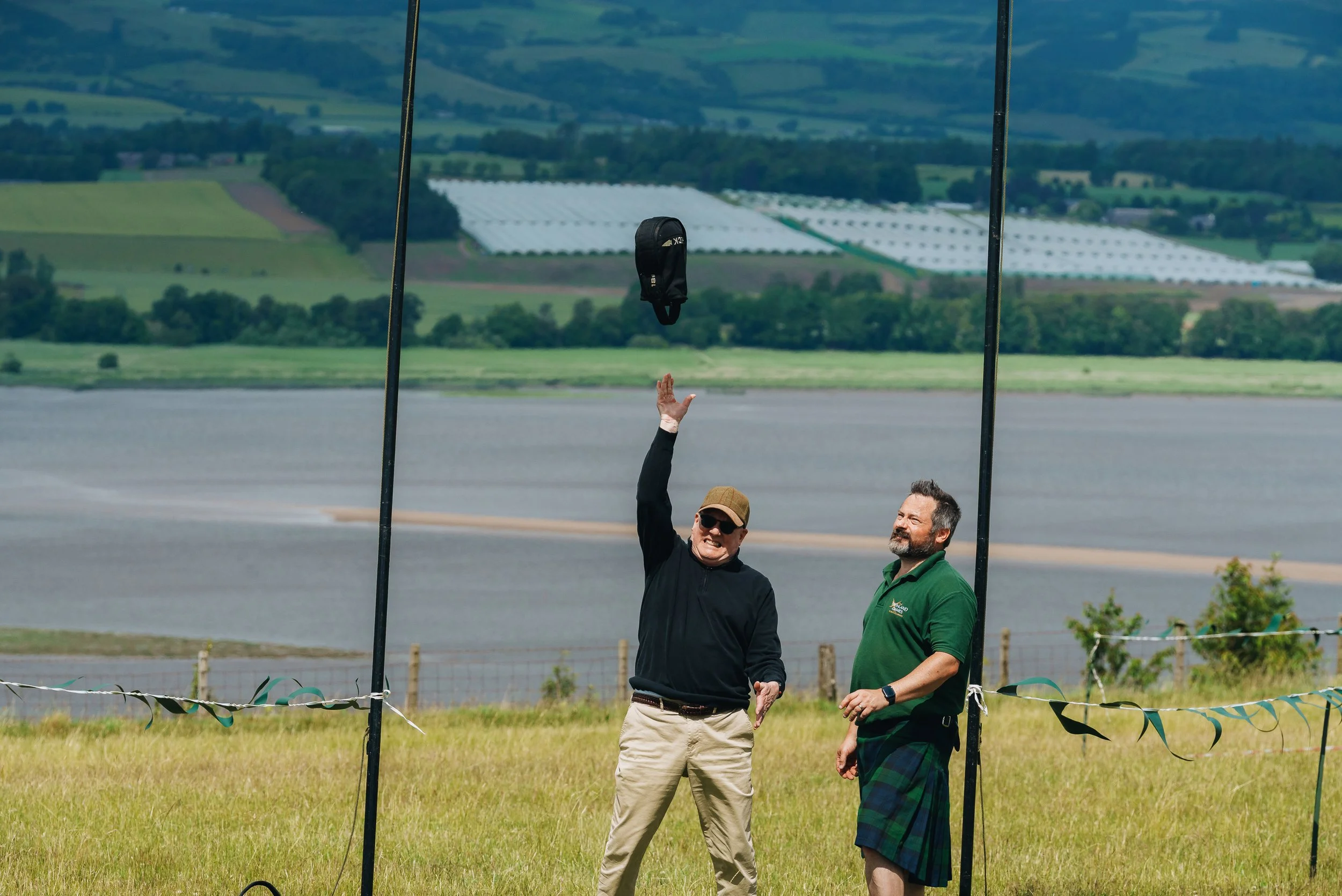 Two men standing on a grassy field near water, with a large harness or frame structure and a bag hanging from it. One man is wearing a black jacket and light-colored pants, the other is in a green shirt and plaid shorts. The look of excitement or ach