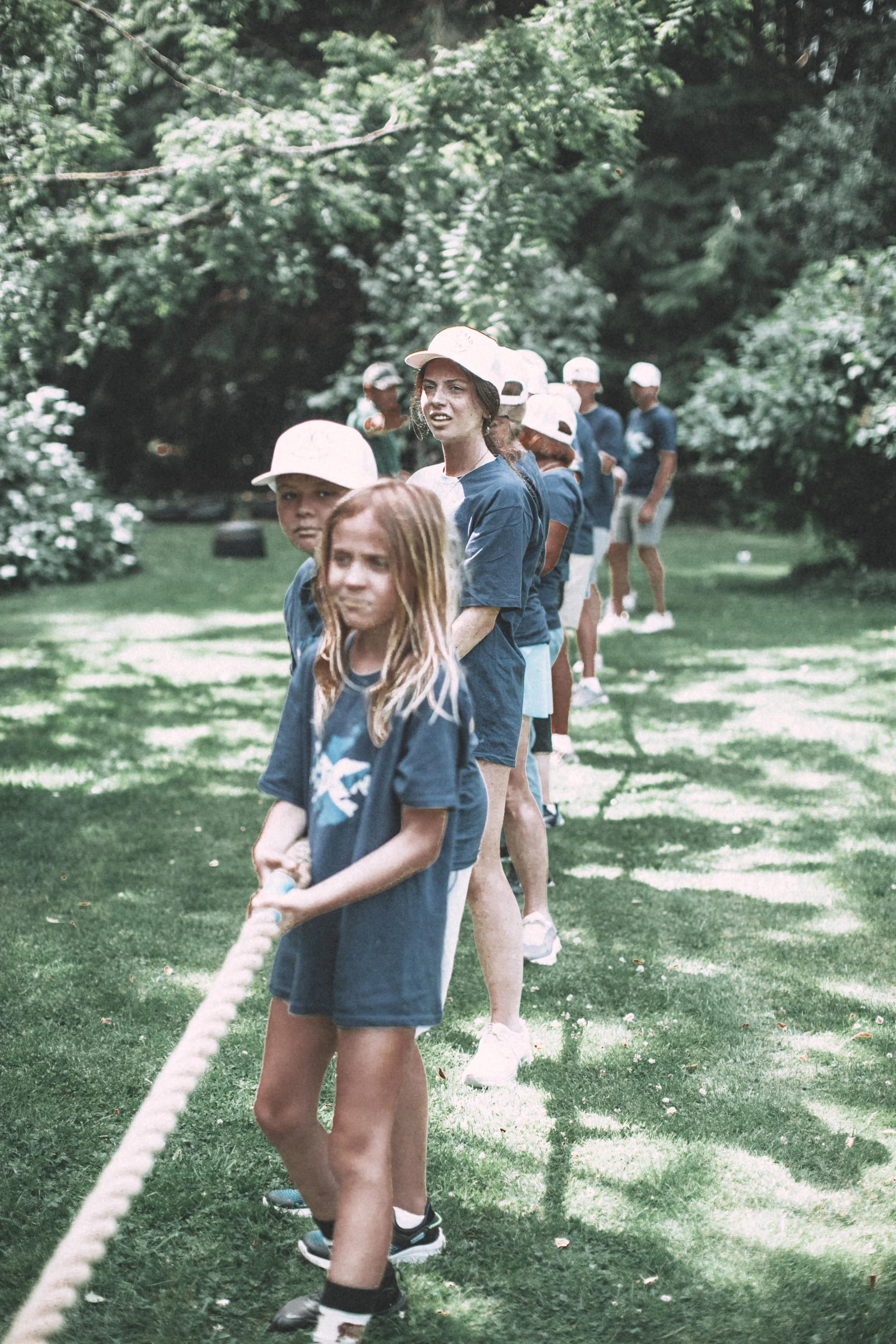 A group of children standing in a line outdoors on a grassy area, holding a rope, with trees and greenery in the background.
