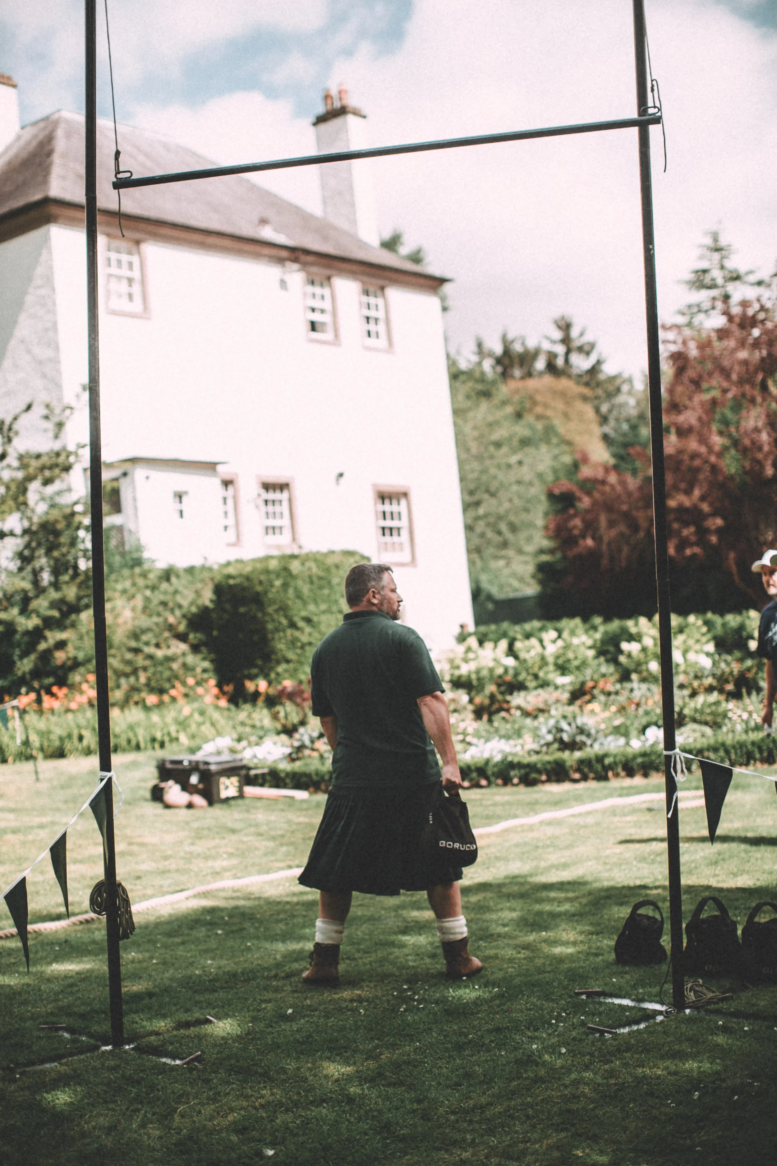 A man dressed in a traditional Scottish kilt stands on a lawn holding a bagpipe case, with tents and a historic house in the background, during an outdoor event.