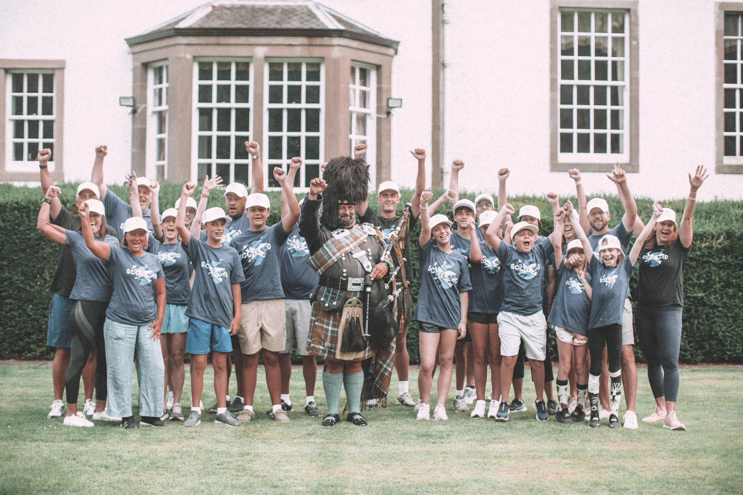 A large group of young people in matching dark blue t-shirts and white caps gathered outdoors on a green lawn, raising their fists in celebration. A person dressed in traditional Scottish Highland dress stands at the front, with a historic building w