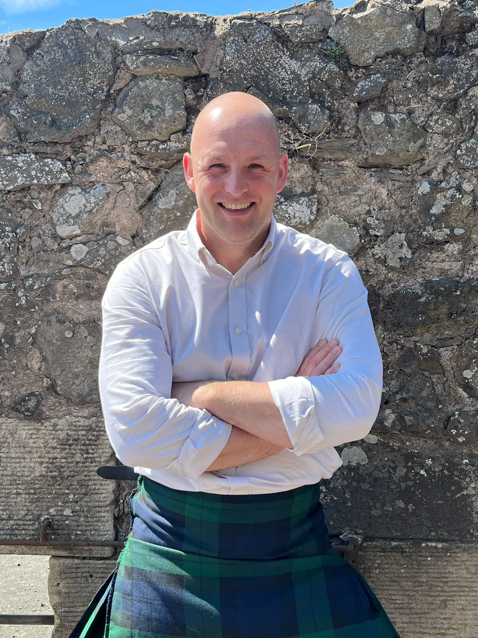 A smiling man with a shaved head, wearing a white dress shirt with rolled-up sleeves and a green and blue tartan kilt, standing with arms crossed in front of a stone wall outdoors.