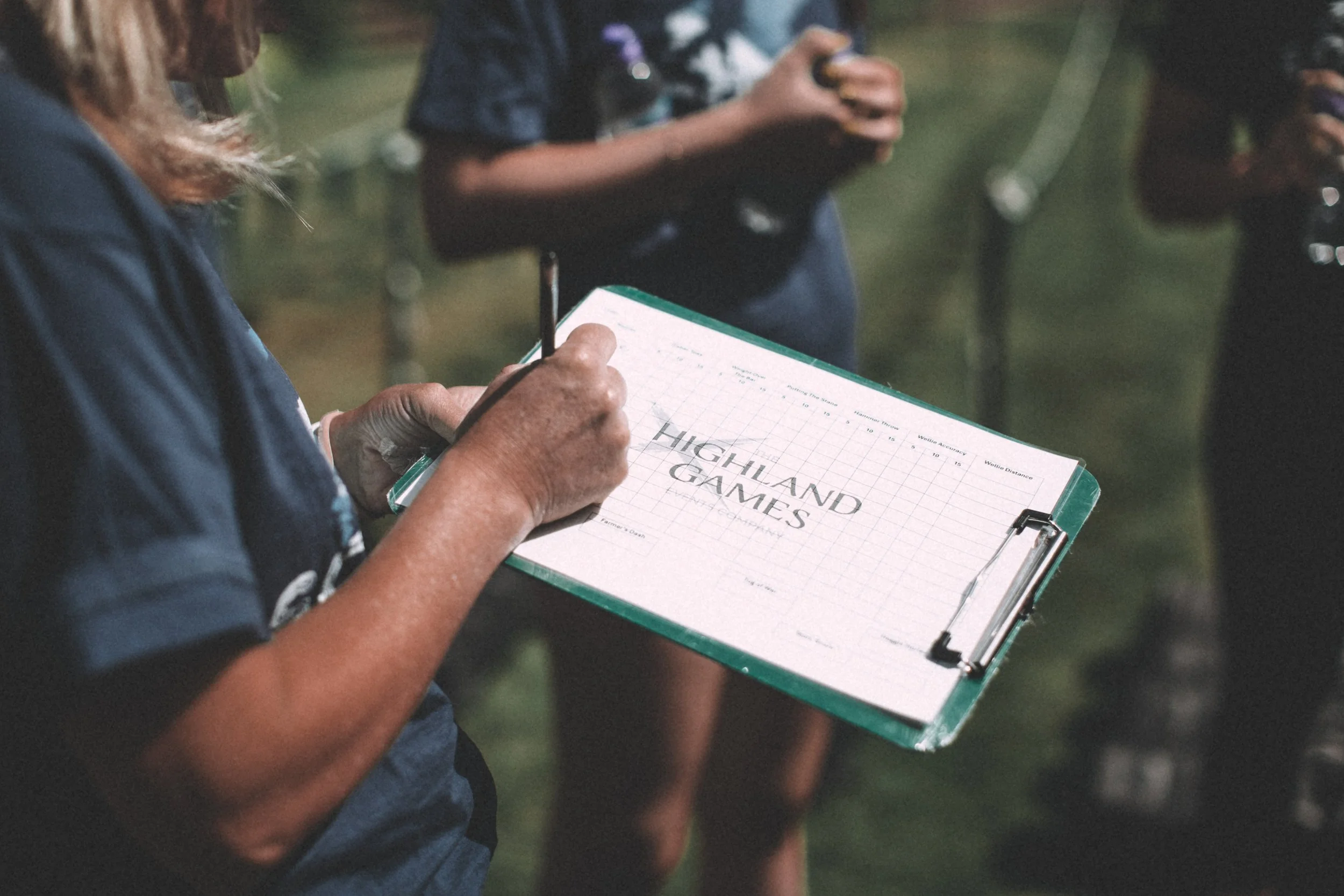 Person in a dark shirt writing on a clipboard that says 'Highland Games' with a pen, while other people are in the background holding tablets or phones.