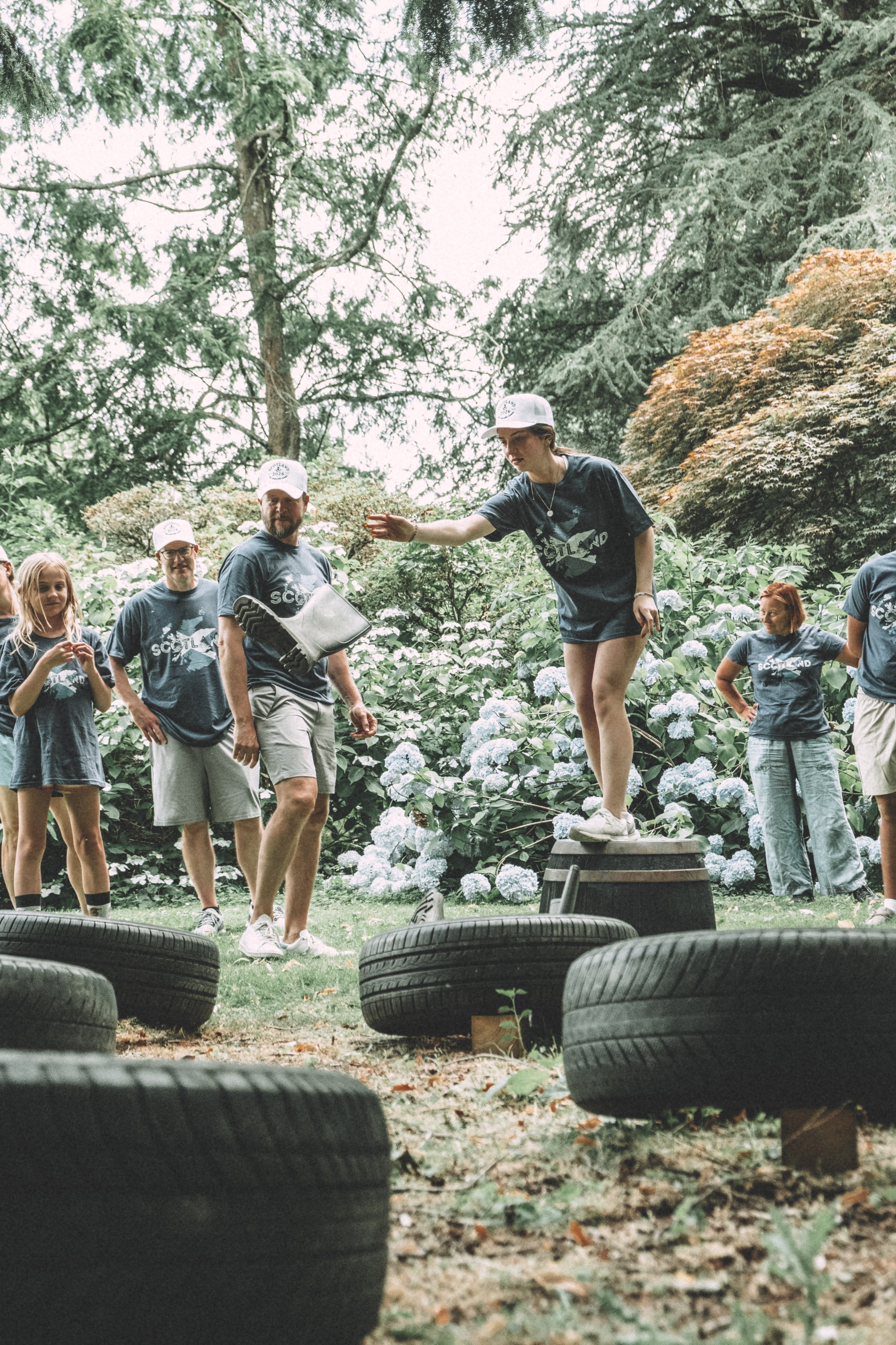 A group of people outdoors in a wooded area, with tires and wooden logs in the foreground, participating in a team activity or game.