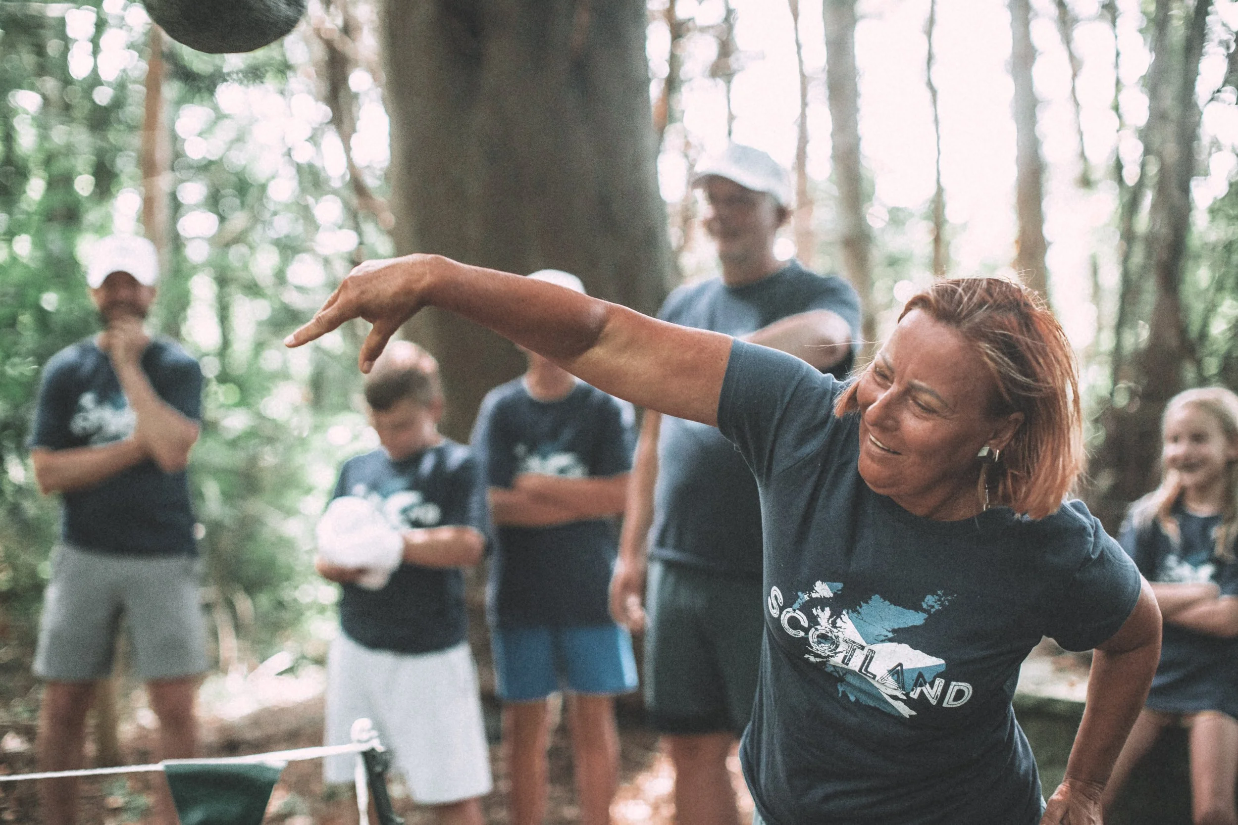 A group of people outdoors in a wooded area, with one woman in the foreground reaching out with her arm, surrounded by others in casual clothing.