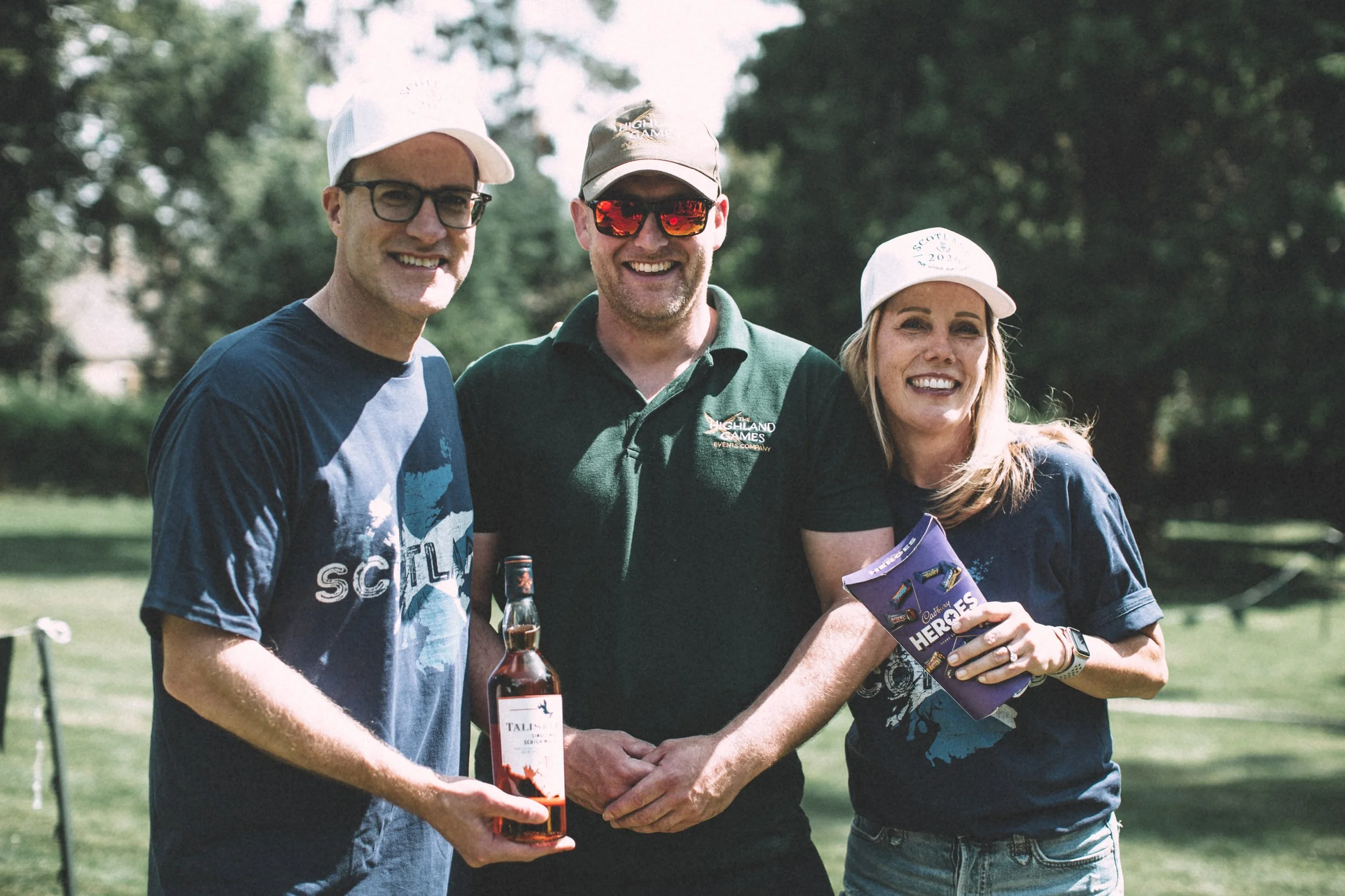 Three smiling people standing outdoors in a park, two men and a woman, holding a bottle of wine. The man on the left is wearing glasses and a white cap, the man in the middle is wearing sunglasses and a cap, and the woman on the right is wearing a wh
