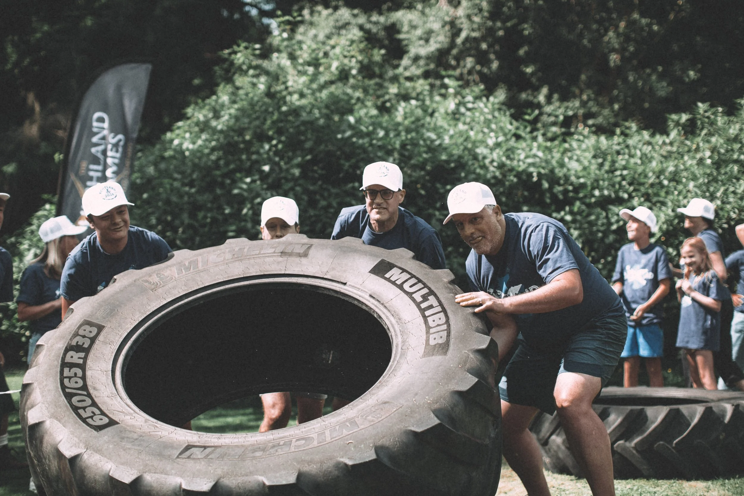 A group of people, including children and adults, participating in a tire flipping activity outdoors with a forested background. The central figures are pushing a large tractor tire.