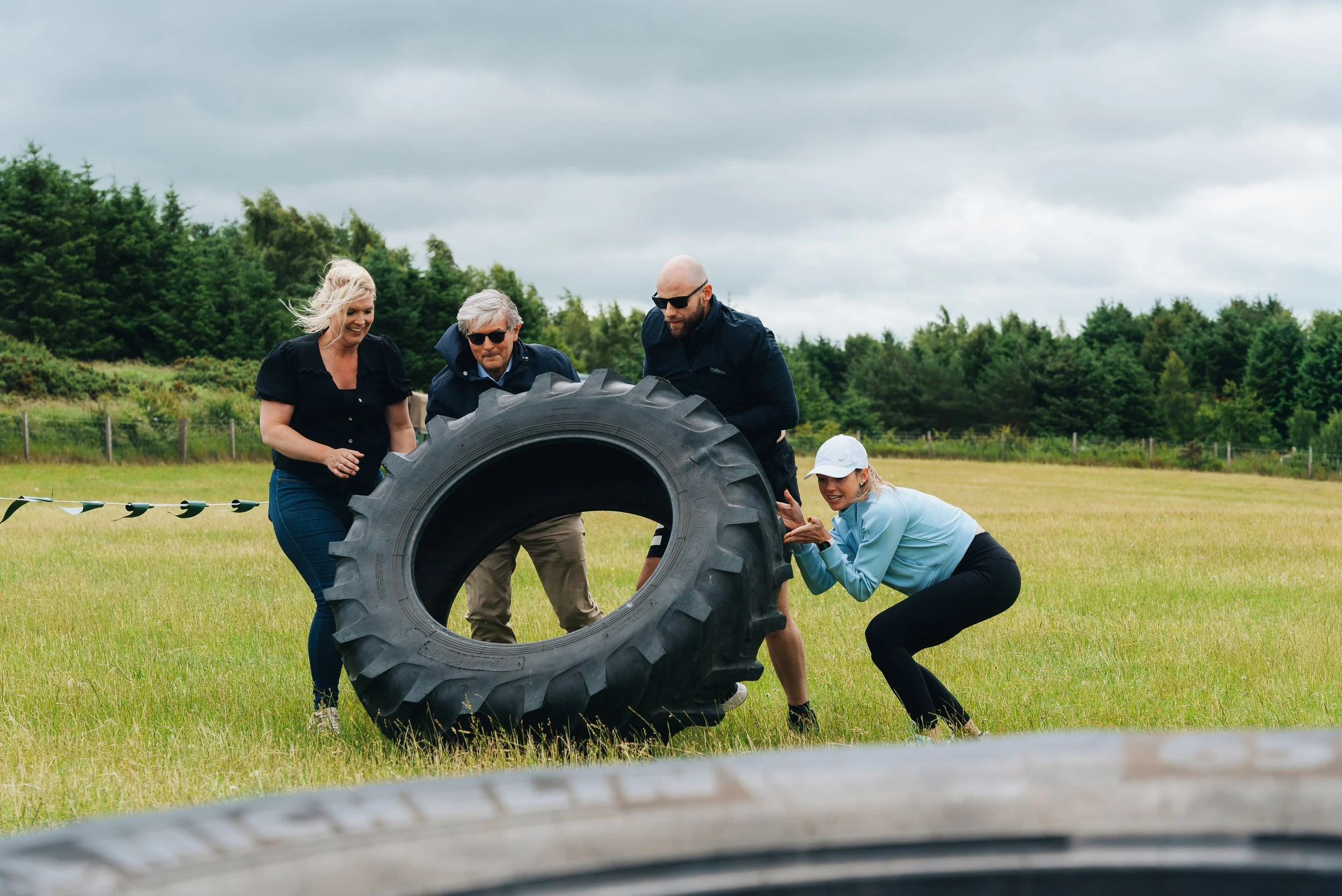 Four people, three adults and one child, working together outdoors to lift a large tractor tire in a grassy field during overcast weather.