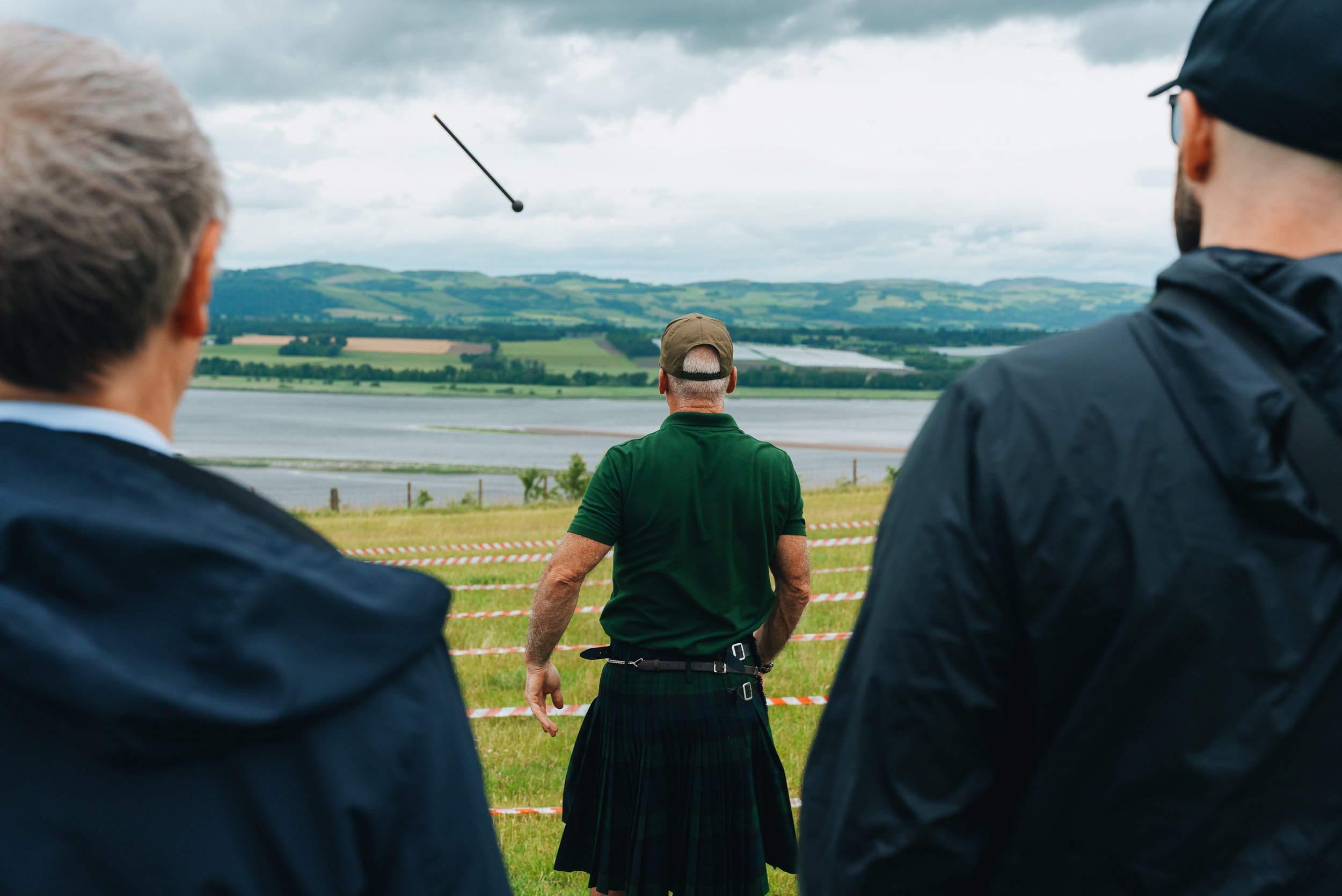 Three men are outdoors near a body of water with rolling hills in the background. The man in the center is wearing a green shirt and a beige cap, and appears to be participating in a golf event. The two men in the foreground are facing away and weari