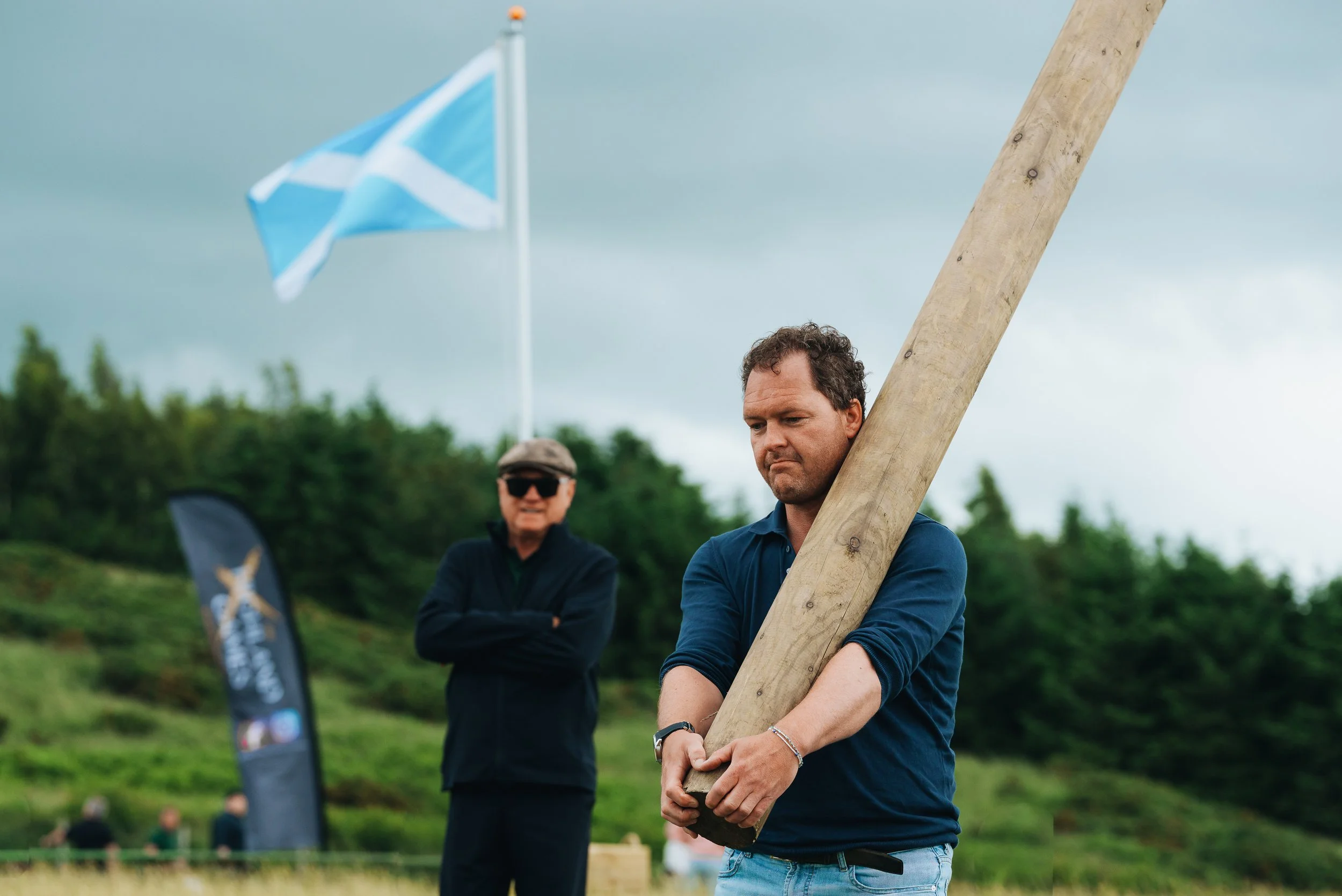 Two men outdoors, one holding a large wooden post, and the other standing with arms crossed. There is a flag in the background and a cloudy sky.