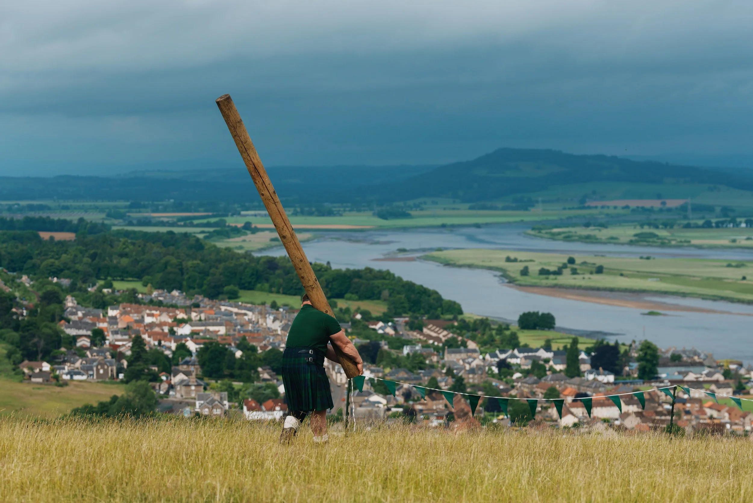 Person holding a traditional wooden staff, standing on a grassy hilltop overlooking a town, river, and rolling hills under cloudy skies.