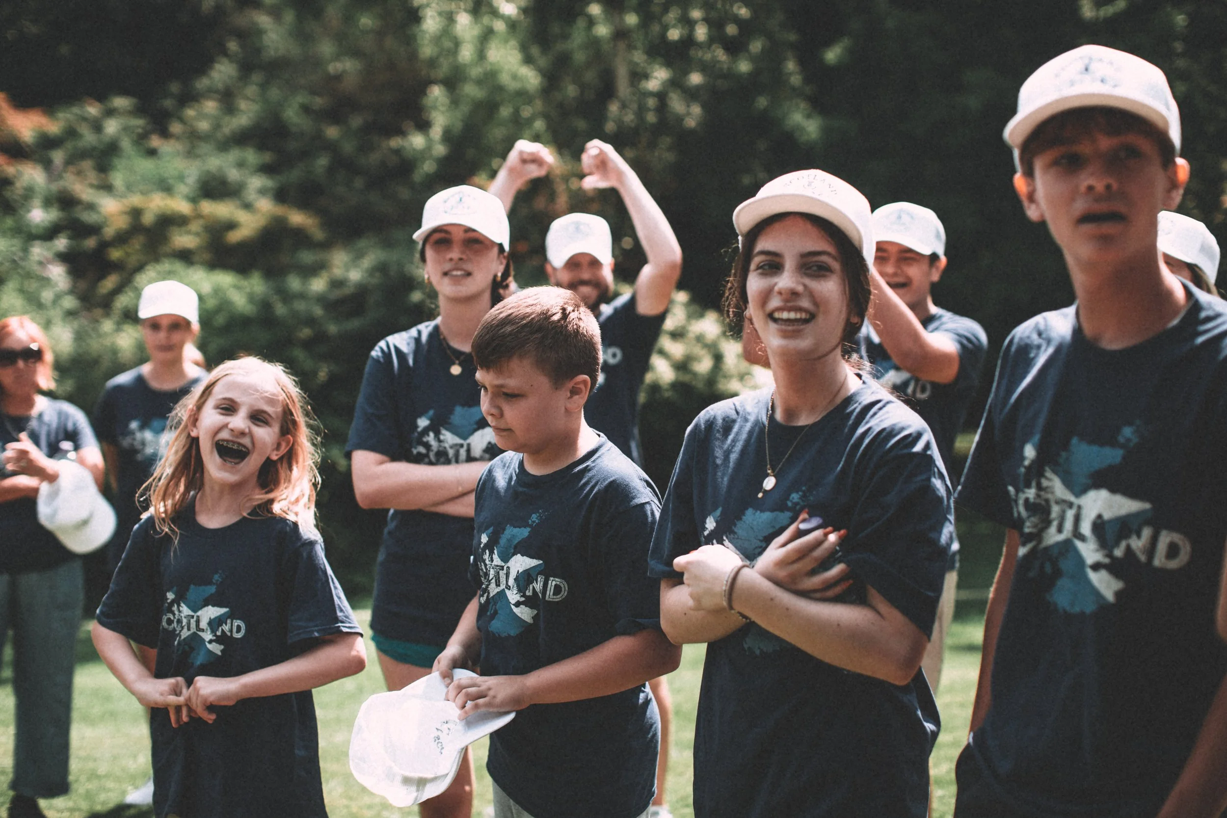 A group of smiling children and teenagers outdoors in a park, wearing matching dark blue t-shirts and white caps, some holding papers, with trees in the background.