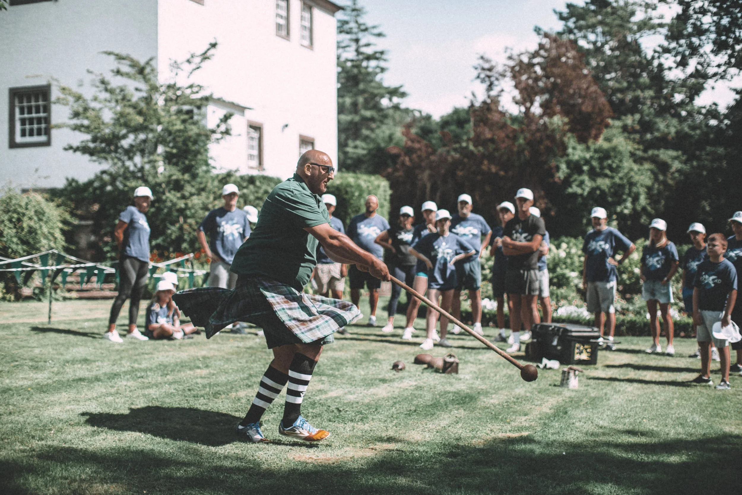 A group of children and a man playing a game outdoors on a field with grass, trees, and a white building in the background. The man is dressed in a kilt and troll grip socks, appears to be in motion, and holding a stick, possibly engaging in a tradit