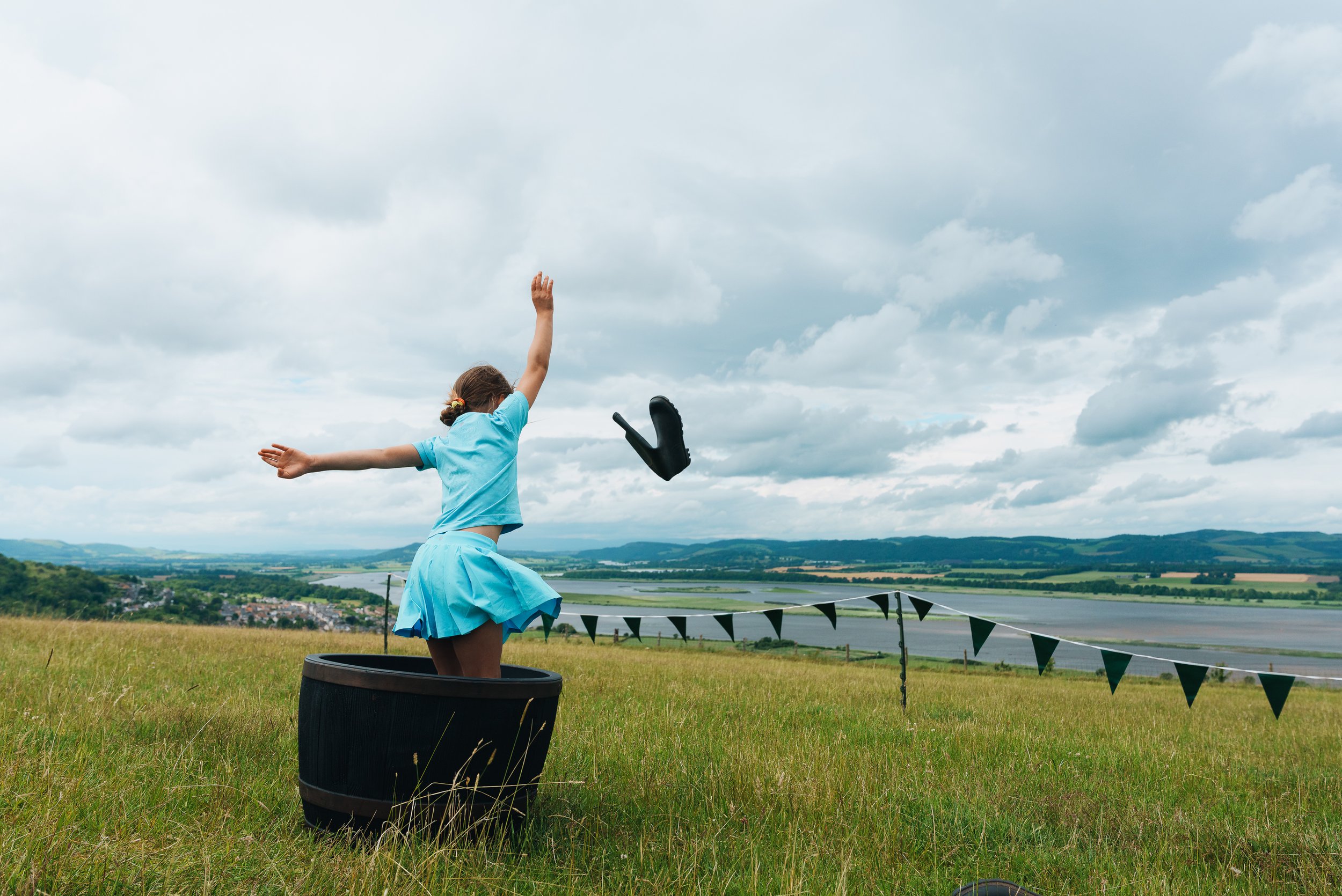 A young girl is jumping into a black barrel with her arms outstretched. She is wearing a blue dress and is outdoors on a grassy hill with a scenic view of water, farmland, and a cloudy sky in the background.