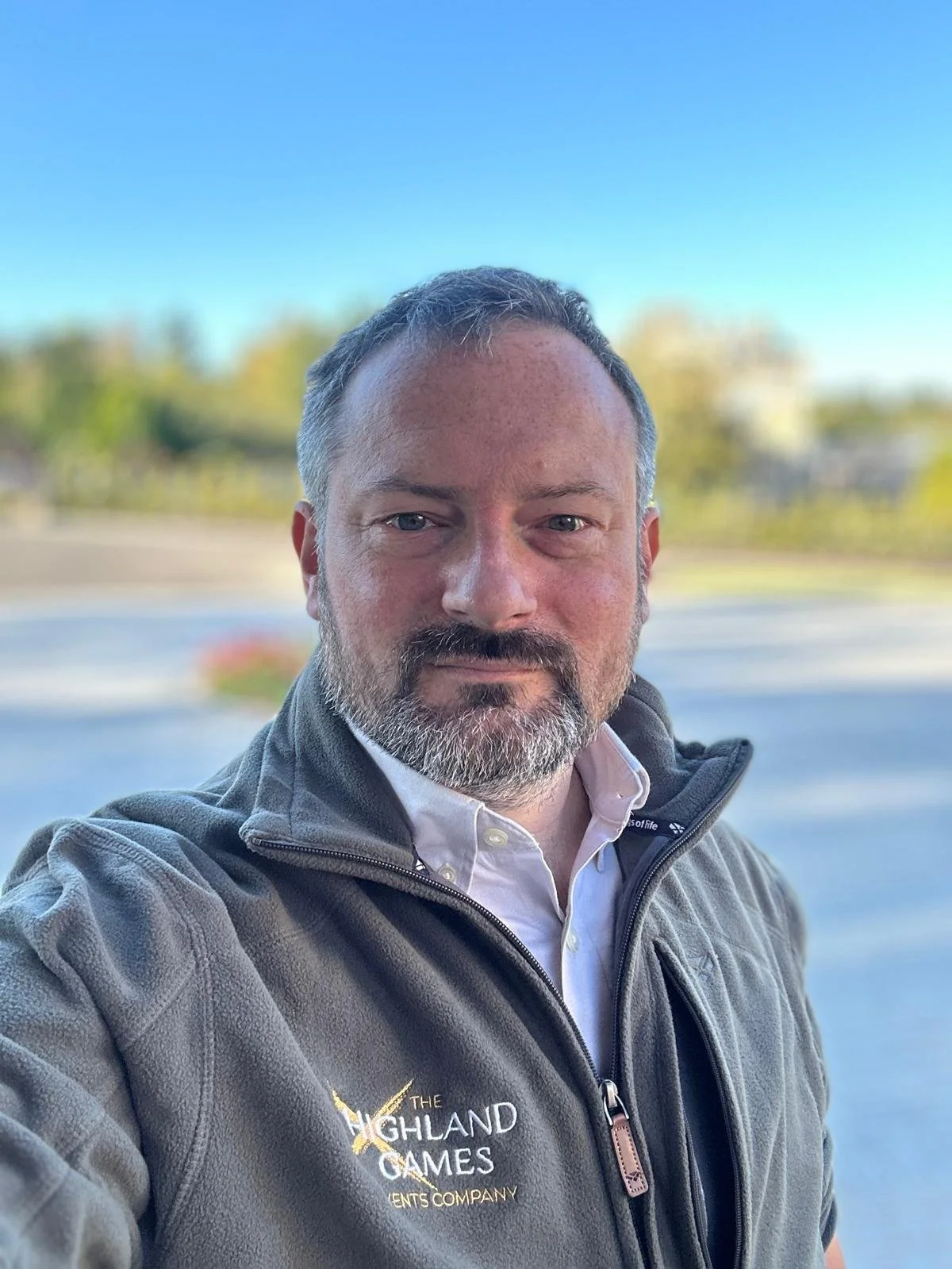 A man taking a selfie outdoors, wearing a gray jacket with the logo 'The Highland Games Events Company,' with a blurred background of trees and a blue sky.
