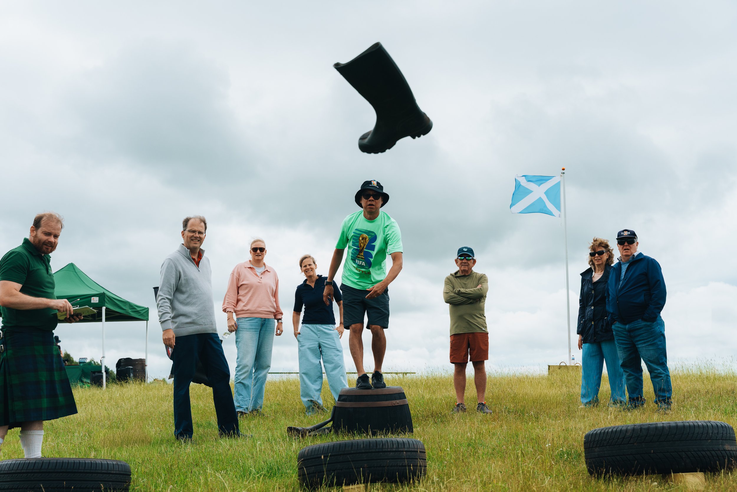 Group of people standing outdoors on a grassy field, participating in a tire-throwing game under cloudy skies, with a Scottish flag in the background.